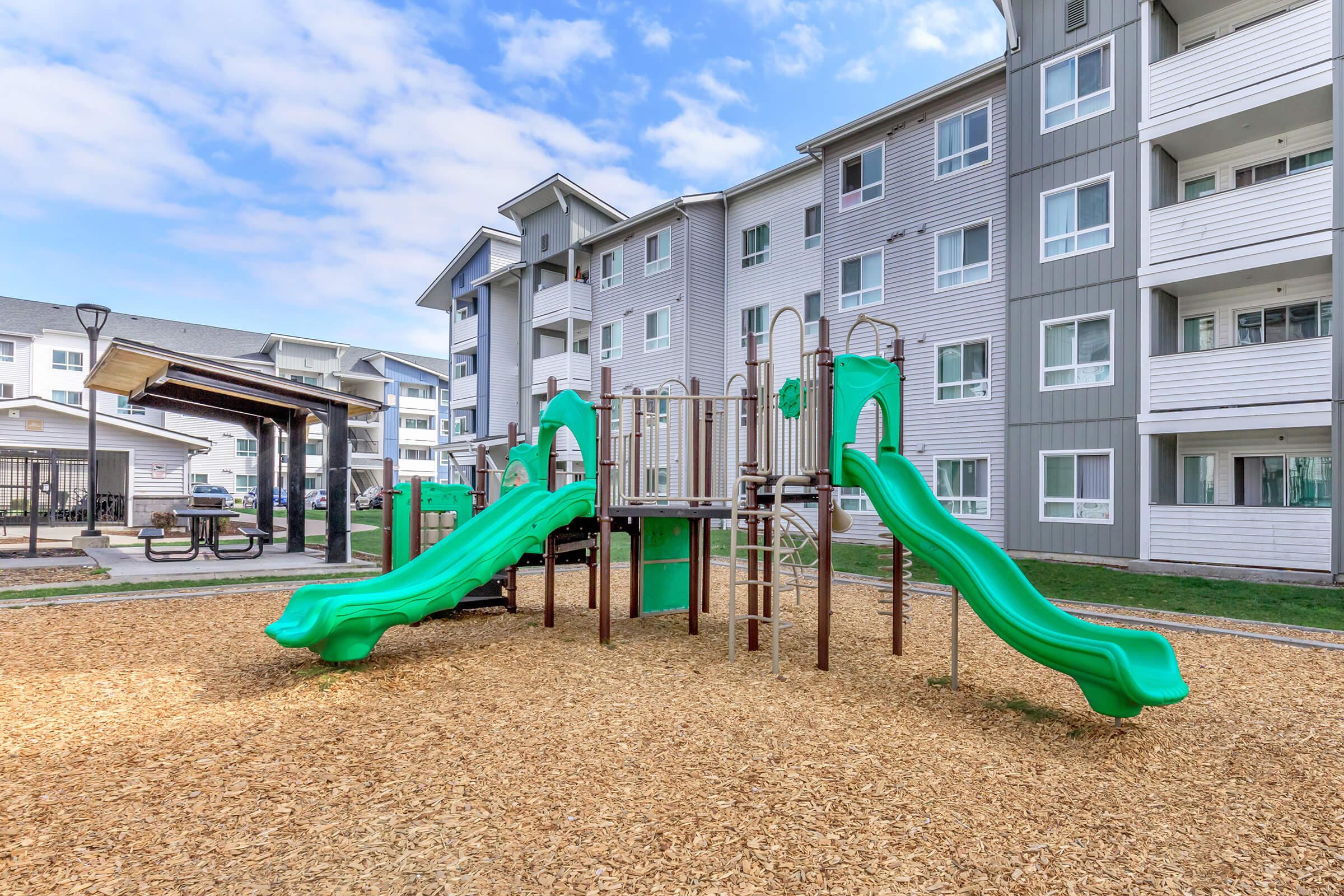 A playground featuring green slides and climbing structures, surrounded by wood chips. In the background, there are multi-story residential buildings with a modern design, under a partly cloudy sky.