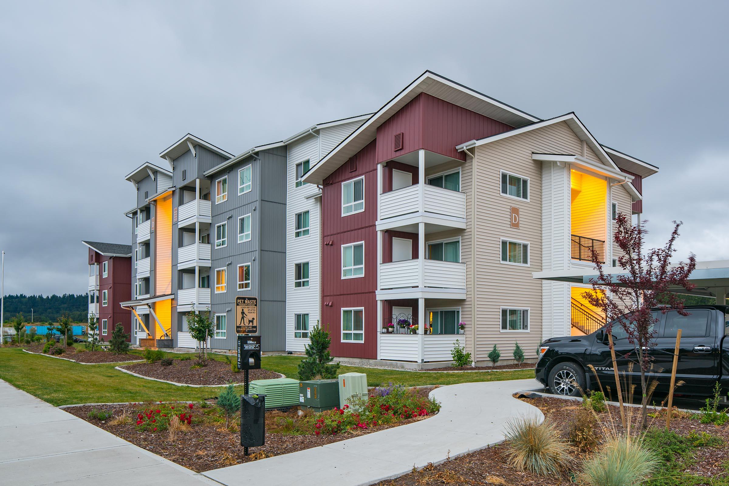 A modern multi-story apartment building featuring a mix of colored facades, with balconies and large windows. The building is surrounded by landscaped greenery, pathways, and utility boxes, under a cloudy sky. A black vehicle is parked nearby.