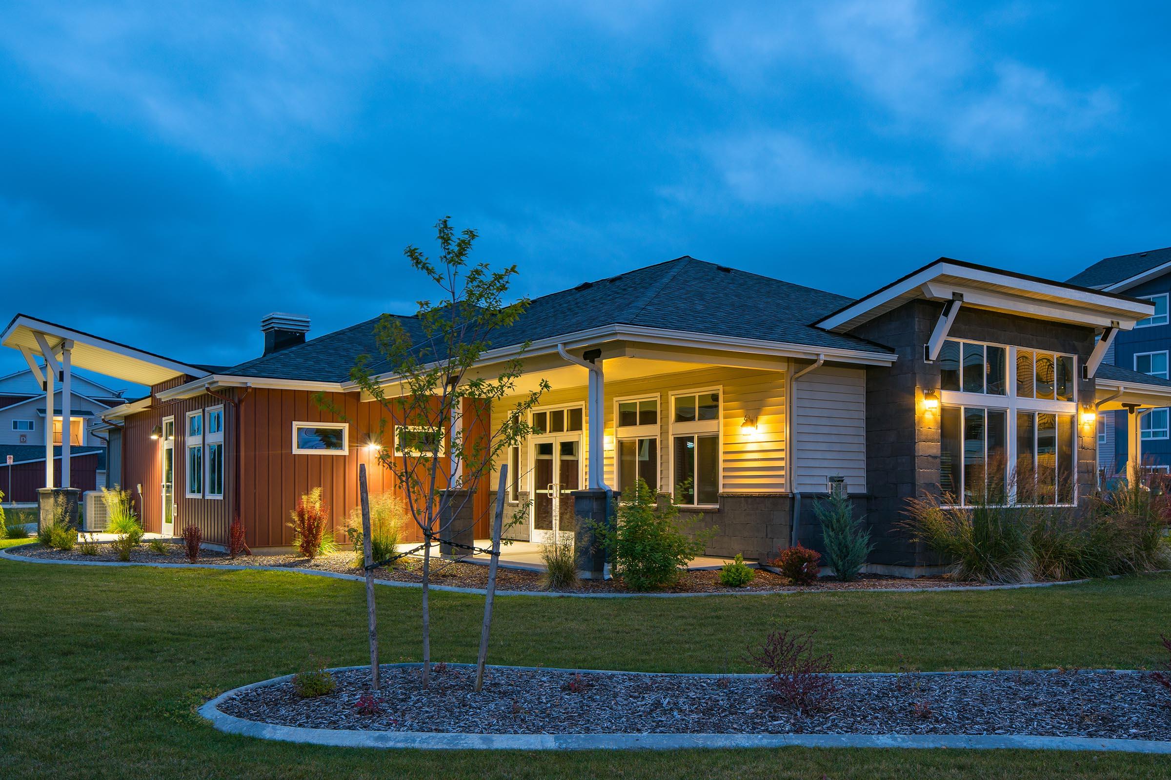 A modern single-story house with a combination of stone and wood exterior, illuminated by warm lights. The home features large windows, a sloped roof, and is surrounded by a landscaped yard with small trees and bushes. The scene is set during twilight with a cloudy sky.