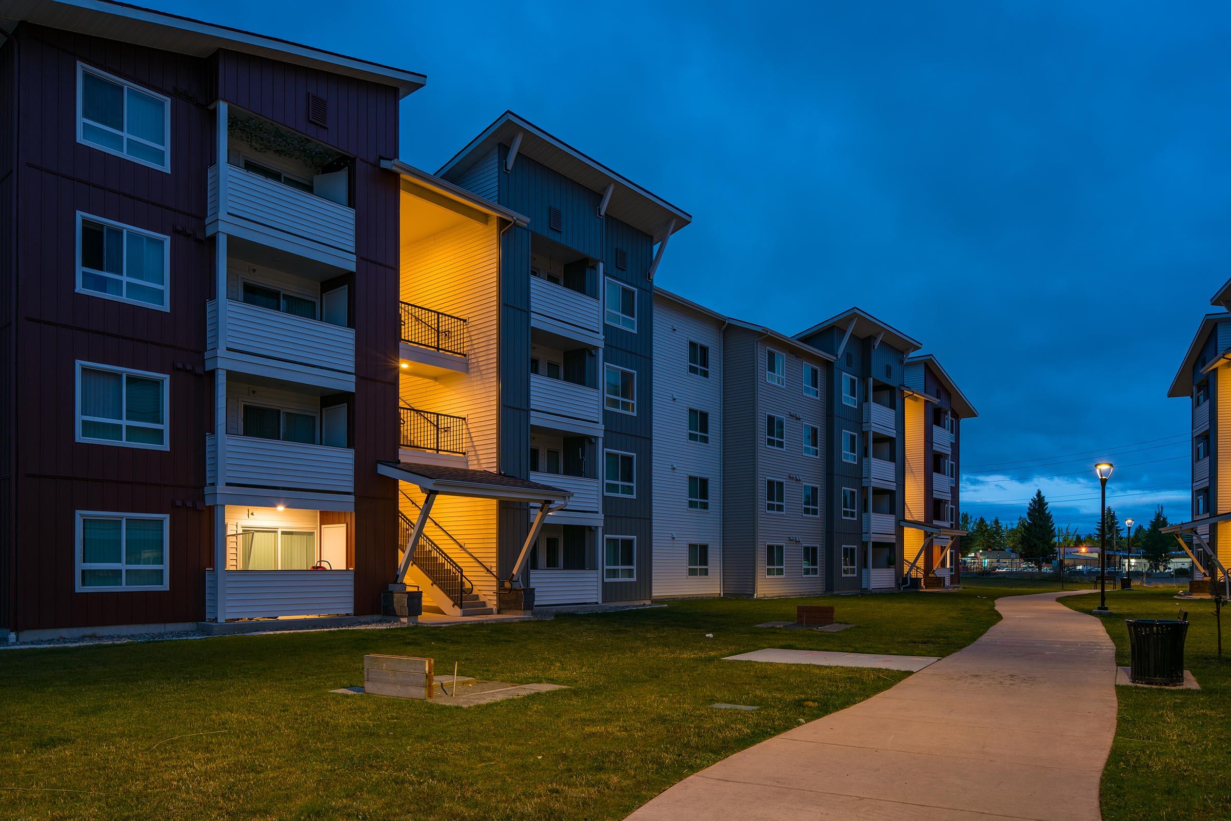 A residential apartment complex at dusk, with several multi-story buildings. One building has a warmly lit entrance, and a pathway leads through a grassy area with a bench. The sky is filled with clouds, creating a serene evening atmosphere.