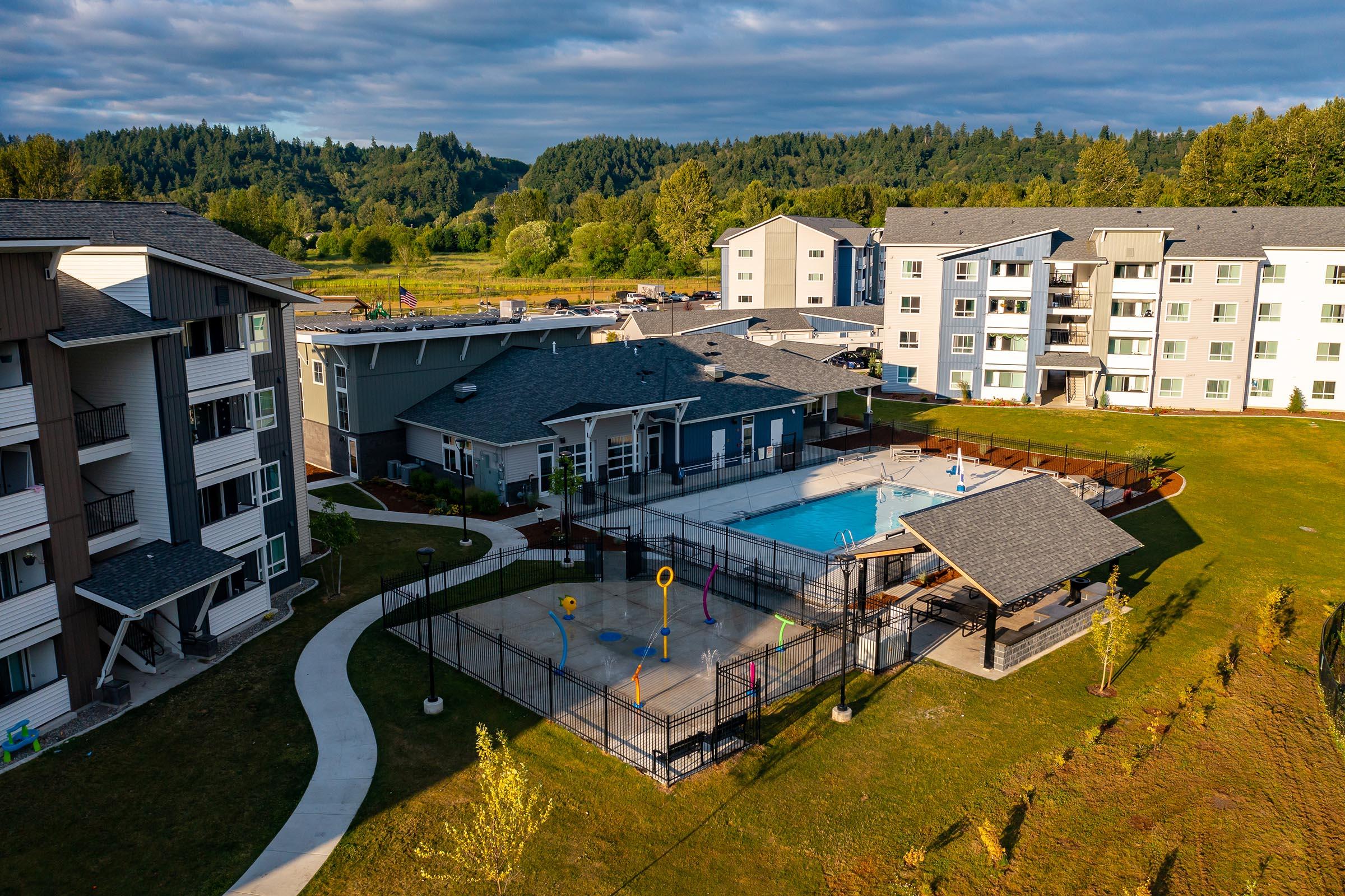 Aerial view of a modern apartment complex with several buildings surrounding a fenced pool area. The pool features colorful play equipment and is situated among green lawns and landscaped gardens. In the background, trees and rolling hills can be seen under a partly cloudy sky.