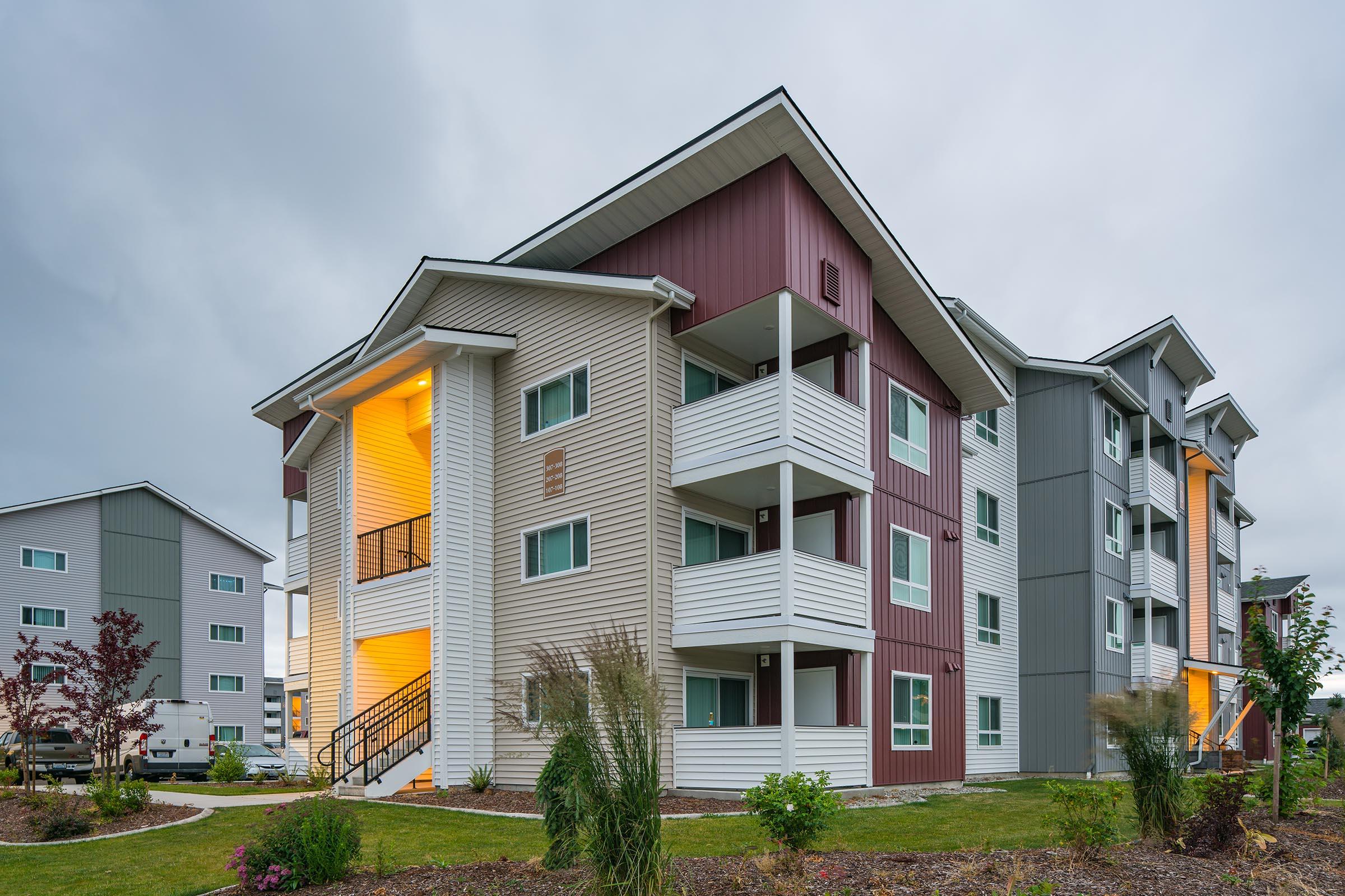 Modern three-story apartment building with a mix of light and dark exterior colors. Features multiple balconies and a landscaped area with ornamental grasses. The setting has a cloudy sky, giving a tranquil atmosphere to the residence.