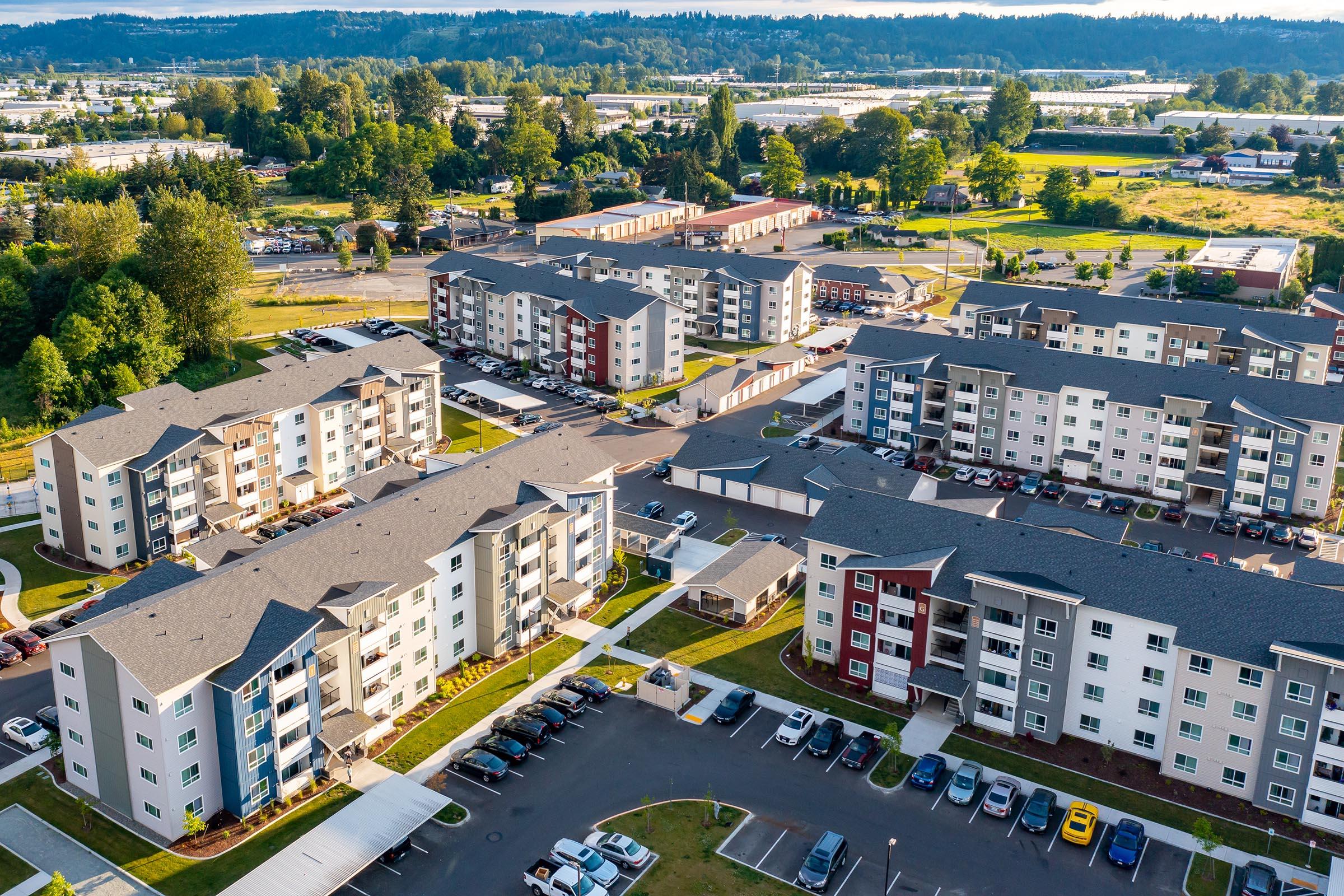 Aerial view of a residential complex featuring multiple apartment buildings with grey and colorful facades. The surrounding area includes green trees and open fields, with parked cars lining the streets and pathways. The scene showcases a mix of urban living and natural landscape.