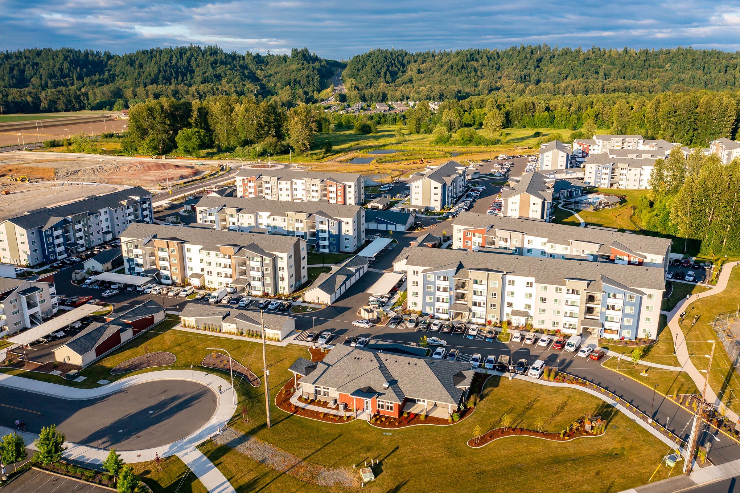 An aerial view of a residential neighborhood featuring multiple apartment buildings surrounded by green areas. The layout includes parking spaces and landscaped grounds, with trees and hills in the background. The scene captures a sunny day with blue skies and scattered clouds.