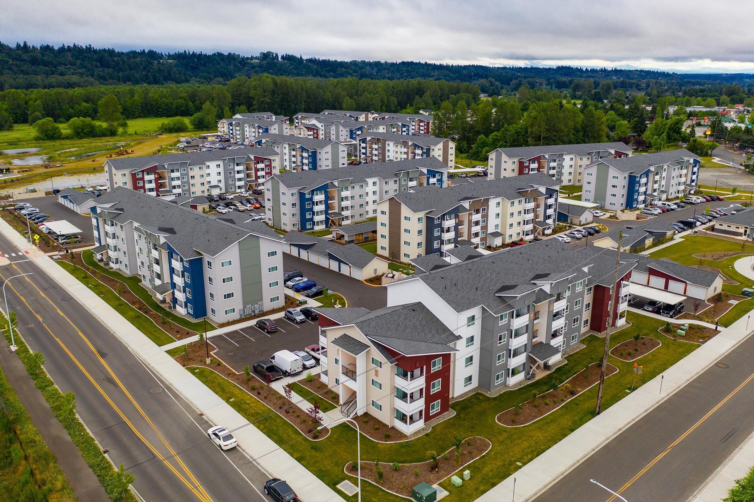 Aerial view of a modern residential complex featuring multiple multi-story buildings with varied color schemes. Surrounding green spaces and roads are visible, along with parked cars. The area appears well-maintained, set against a backdrop of trees and hills under a cloudy sky.