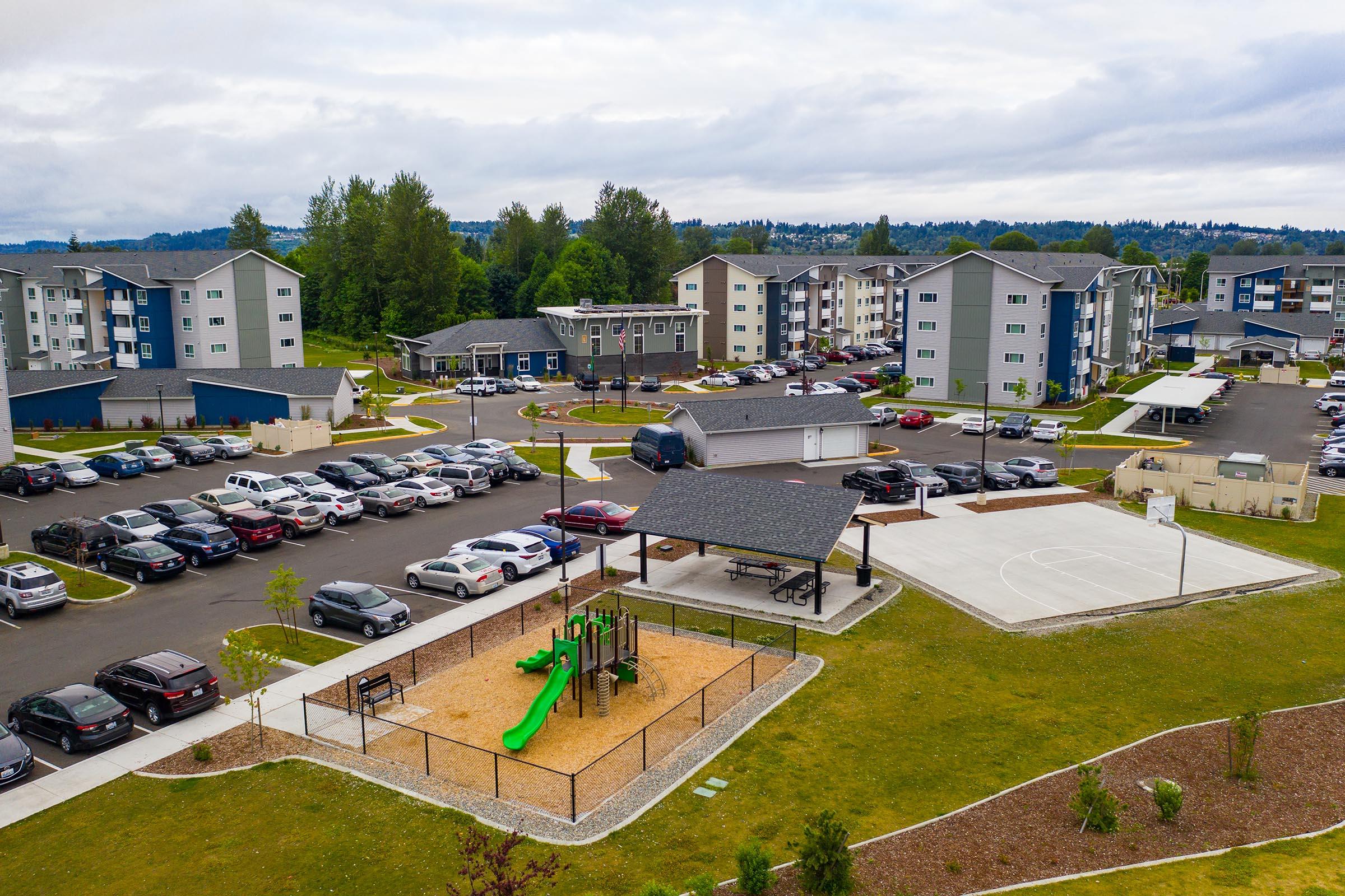Aerial view of a residential area featuring multiple apartment buildings surrounded by a parking lot. In the foreground, there's a playground with a slide, a sandy area, and a basketball court. Lush green grass and trees are visible, along with several parked cars and a covered picnic area.