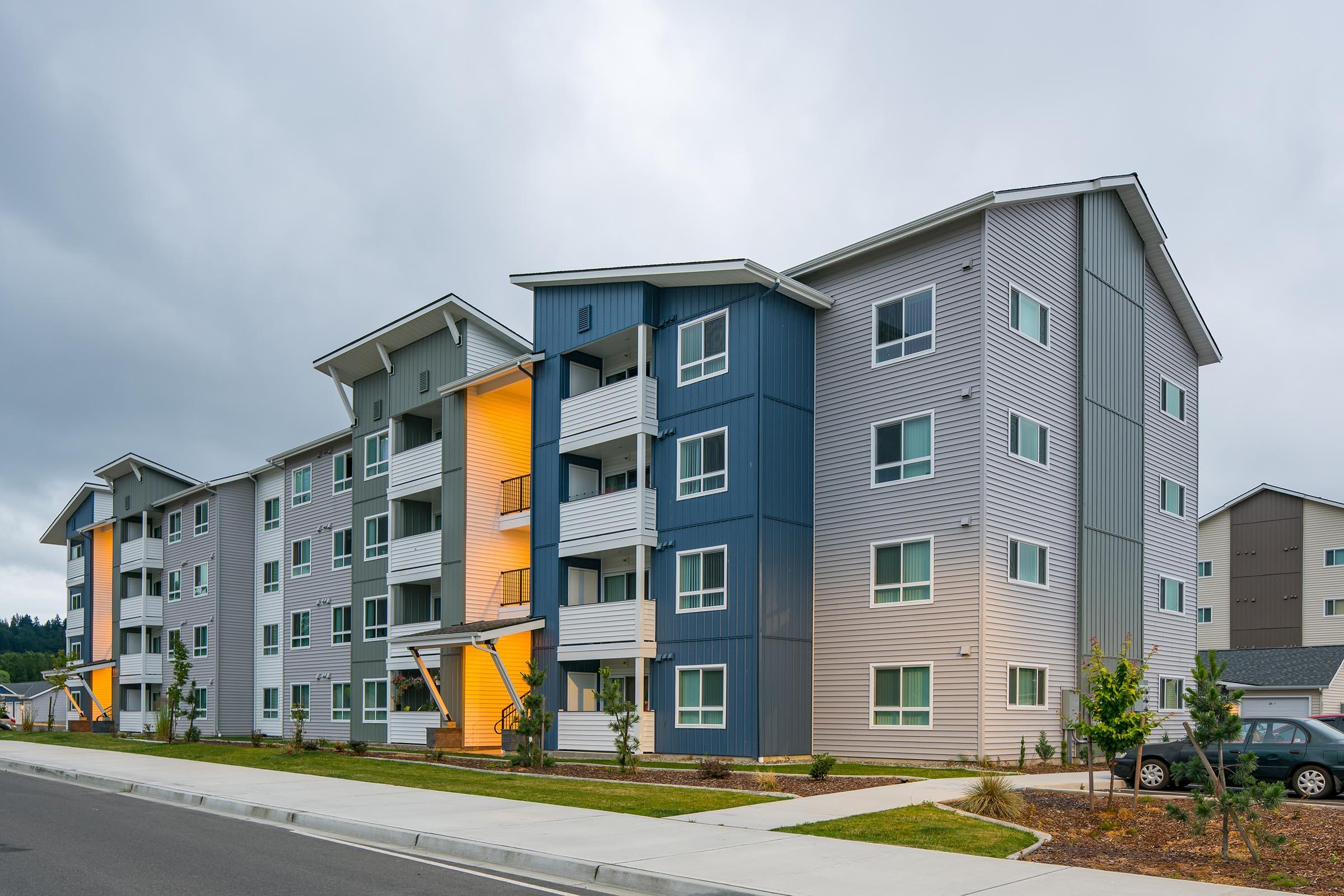 A modern multi-story apartment building featuring a mix of light gray and blue siding with orange accents. The structure has multiple balconies and large windows, surrounded by a landscaped area. The scene is set on a cloudy day with a wide street in the foreground.