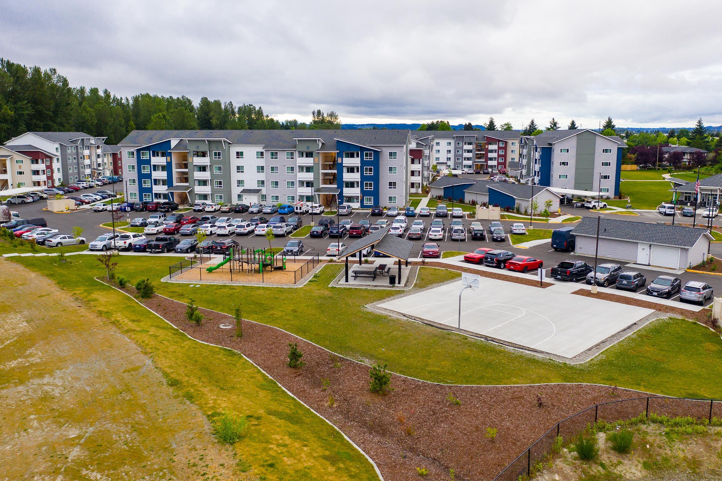 Aerial view of a residential complex with multiple buildings and parking lots. In the foreground, there is a playground area and a basketball court. The surrounding landscape features green lawns and trees, with cloudy skies in the background.