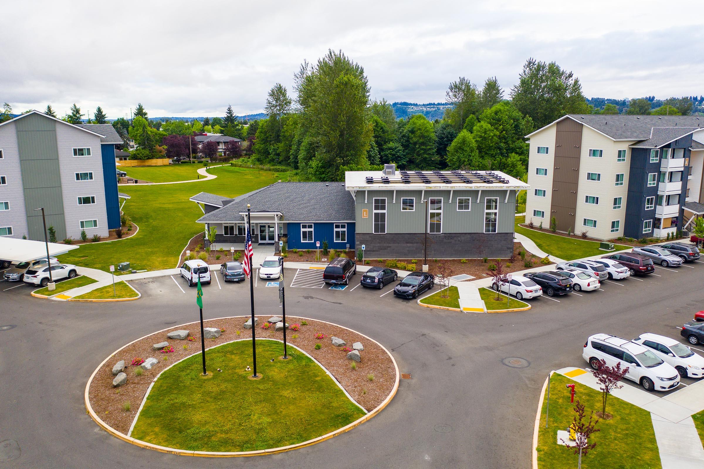Aerial view of a residential complex featuring multiple buildings surrounded by green lawns and trees. In the foreground, a circular driveway includes several parked cars and flagpoles with American and state flags. The sky is cloudy, creating a serene atmosphere.