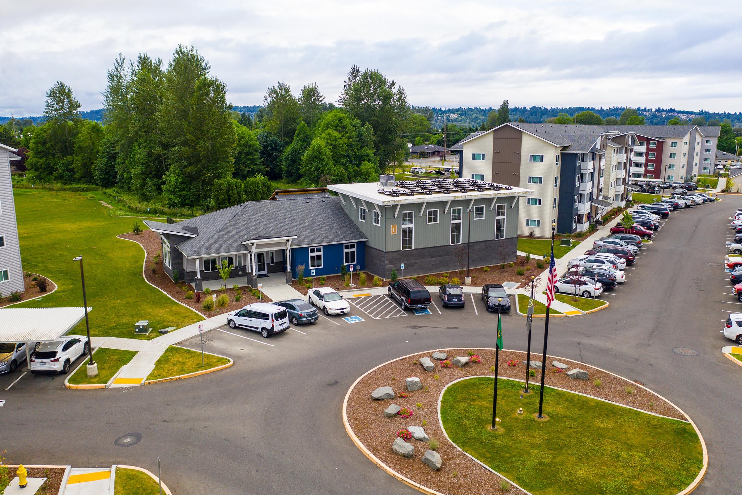 Aerial view of a residential community featuring modern apartment buildings, a landscaped area with trees and shrubs, a circular driveway with parked cars, and a clubhouse or office building. Flagpoles display flags, and there are walking paths and green spaces around the complex.