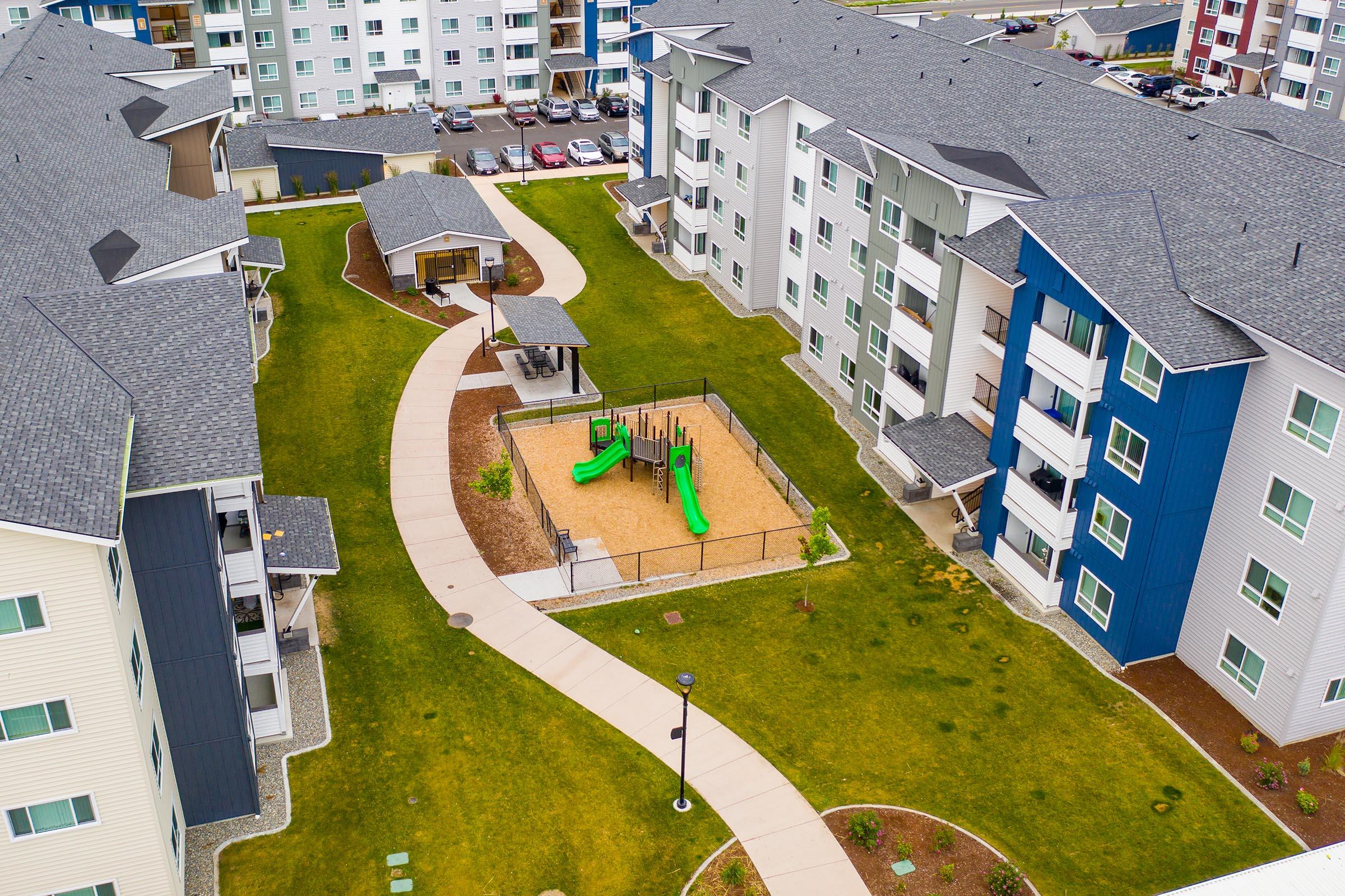 Aerial view of a residential complex featuring multiple buildings with gray roofs. In the center, a landscaped area includes a playground with green slides and a sandbox, surrounded by grassy paths. Several parked cars are visible nearby, along with outdoor seating and walking paths.