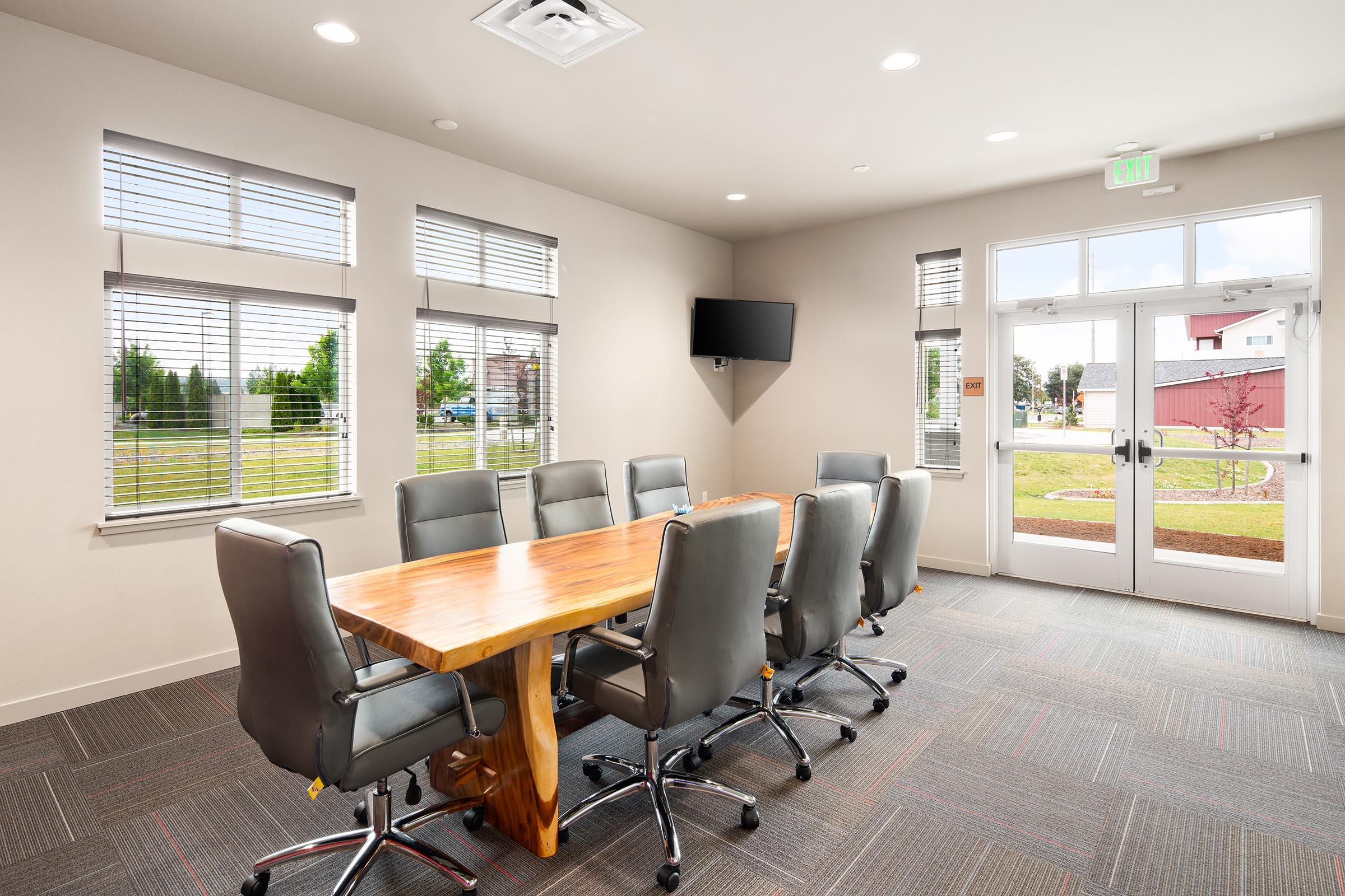 A modern conference room featuring a large wooden table surrounded by gray executive chairs. Natural light enters through several windows, and a flat-screen TV is mounted on the wall. The room has a neutral color palette with carpeted flooring and a door leading outside.