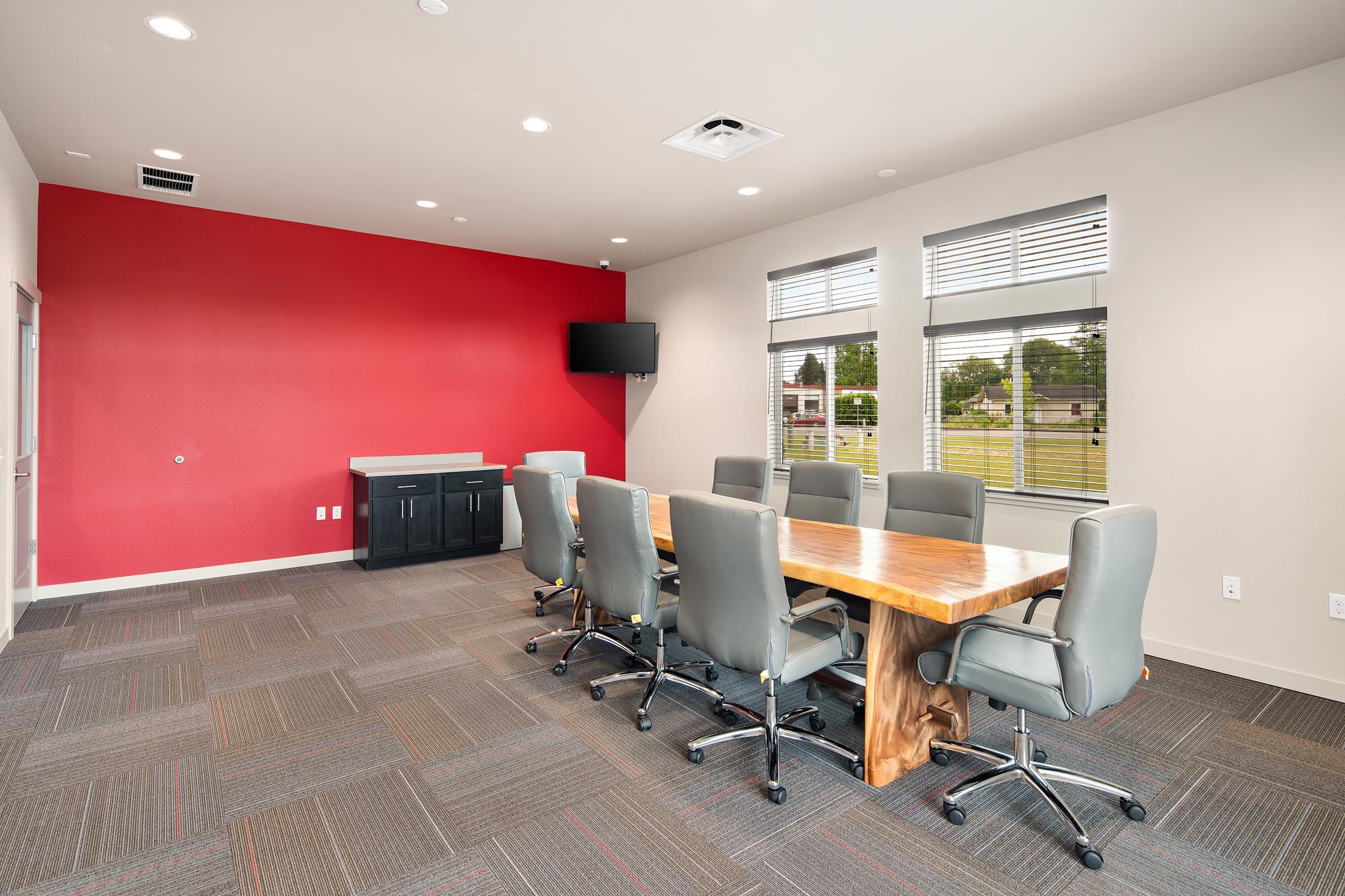 A modern conference room featuring a large wooden table surrounded by six gray office chairs. A red accent wall contrasts with neutral-colored walls. There is a flat-screen TV mounted on the wall and a side cabinet in the background. Natural light floods in through several windows.