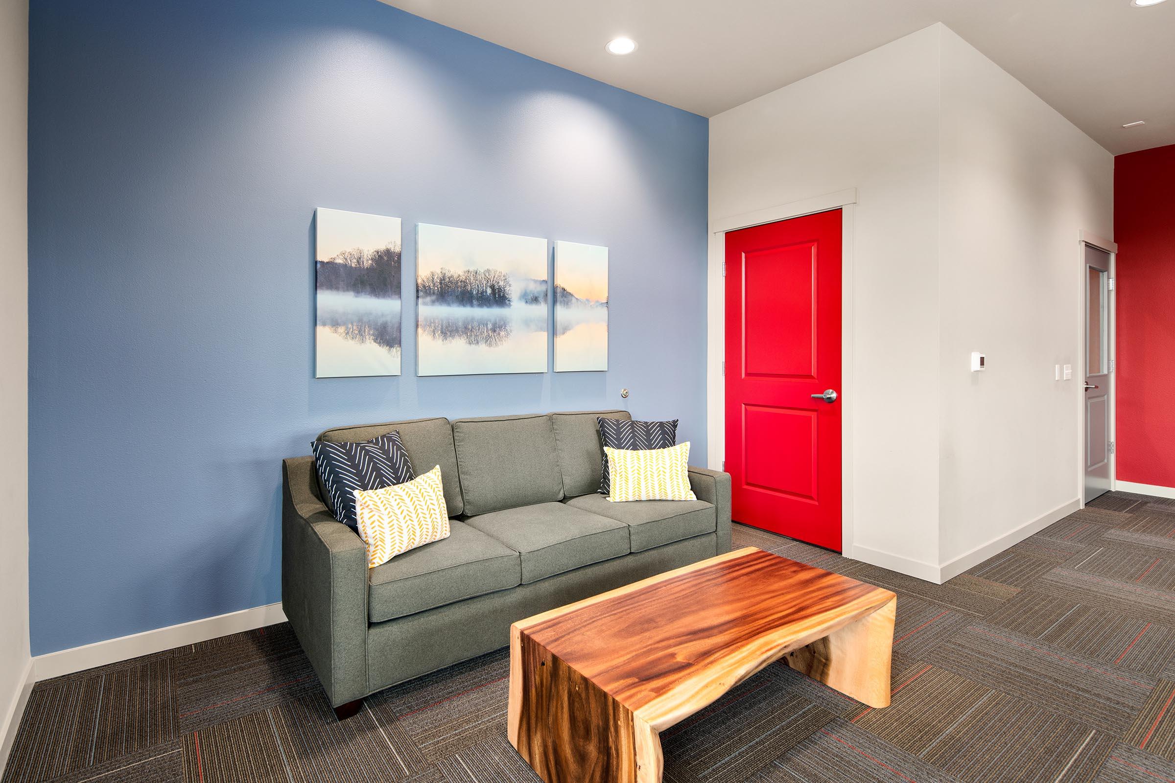 A cozy waiting area featuring a gray sofa with textured yellow and navy pillows, paired with a natural wood coffee table. The wall behind is painted blue and displays three landscape art pieces of a lake. A bright red door contrasts with the serene color palette of the room.