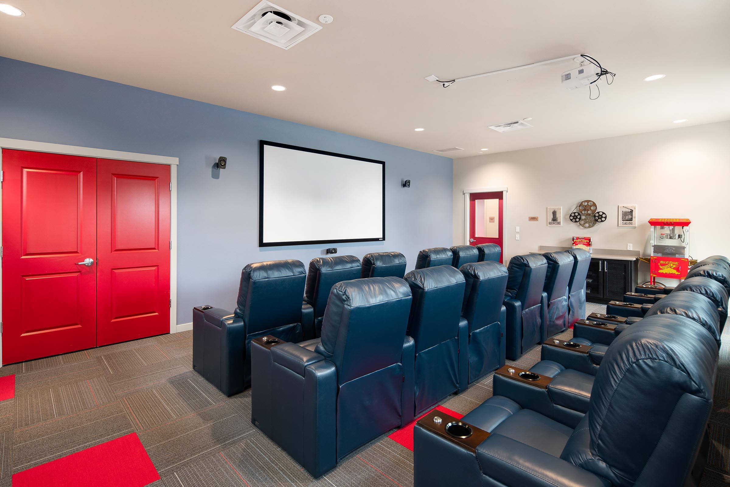 A home theater room featuring blue reclining seats arranged in front of a large screen. The walls are painted in light colors, with a red door and accents. A popcorn machine is visible in the corner, and the space is designed for comfortable viewing and entertainment.