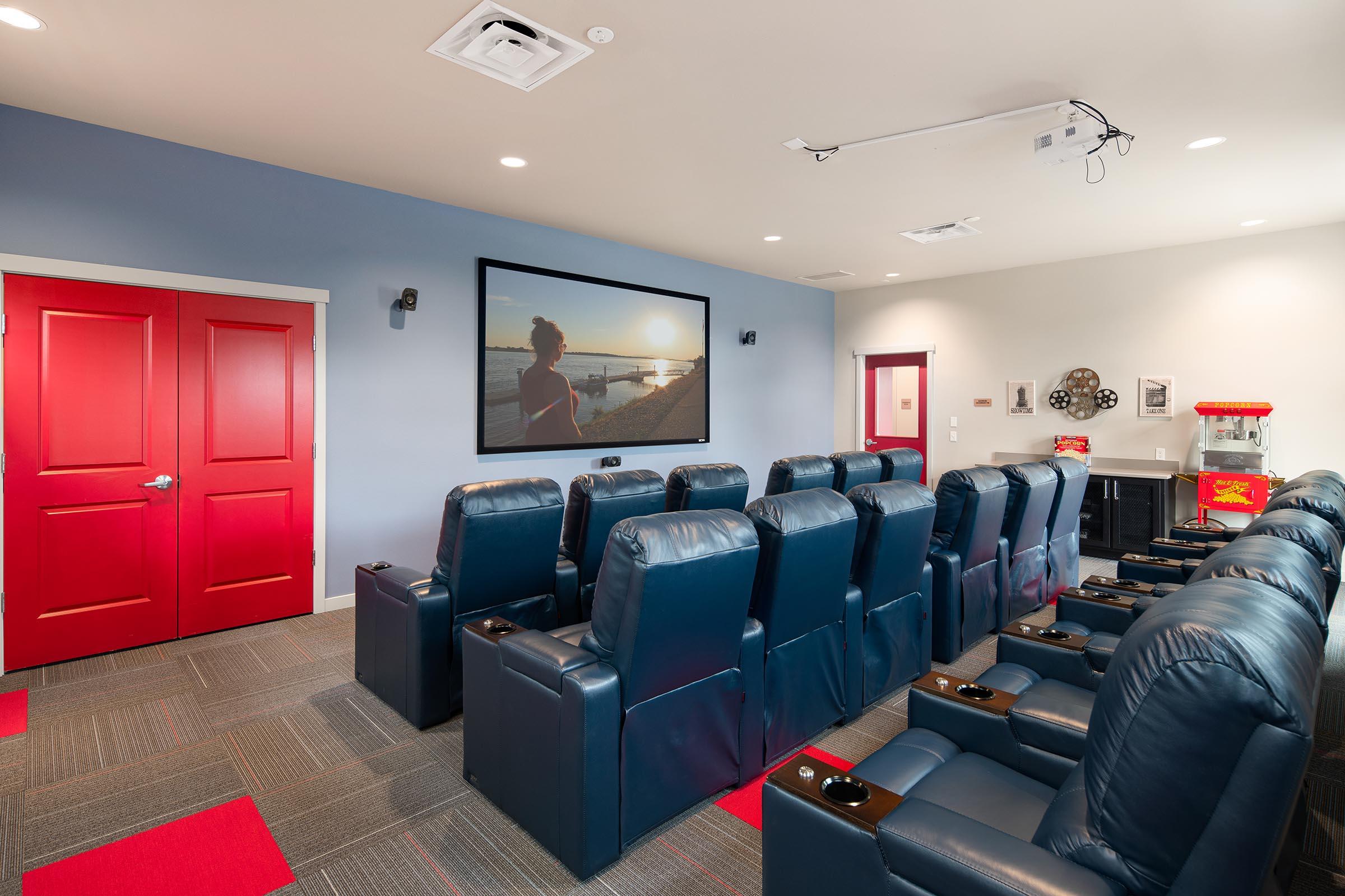Home theater room featuring plush blue reclining chairs arranged in rows, a large screen displaying a sunset scene, red doors, and a popcorn machine in the corner. The room has a modern design with gray and red accents, creating a cozy, inviting atmosphere for movie watching.