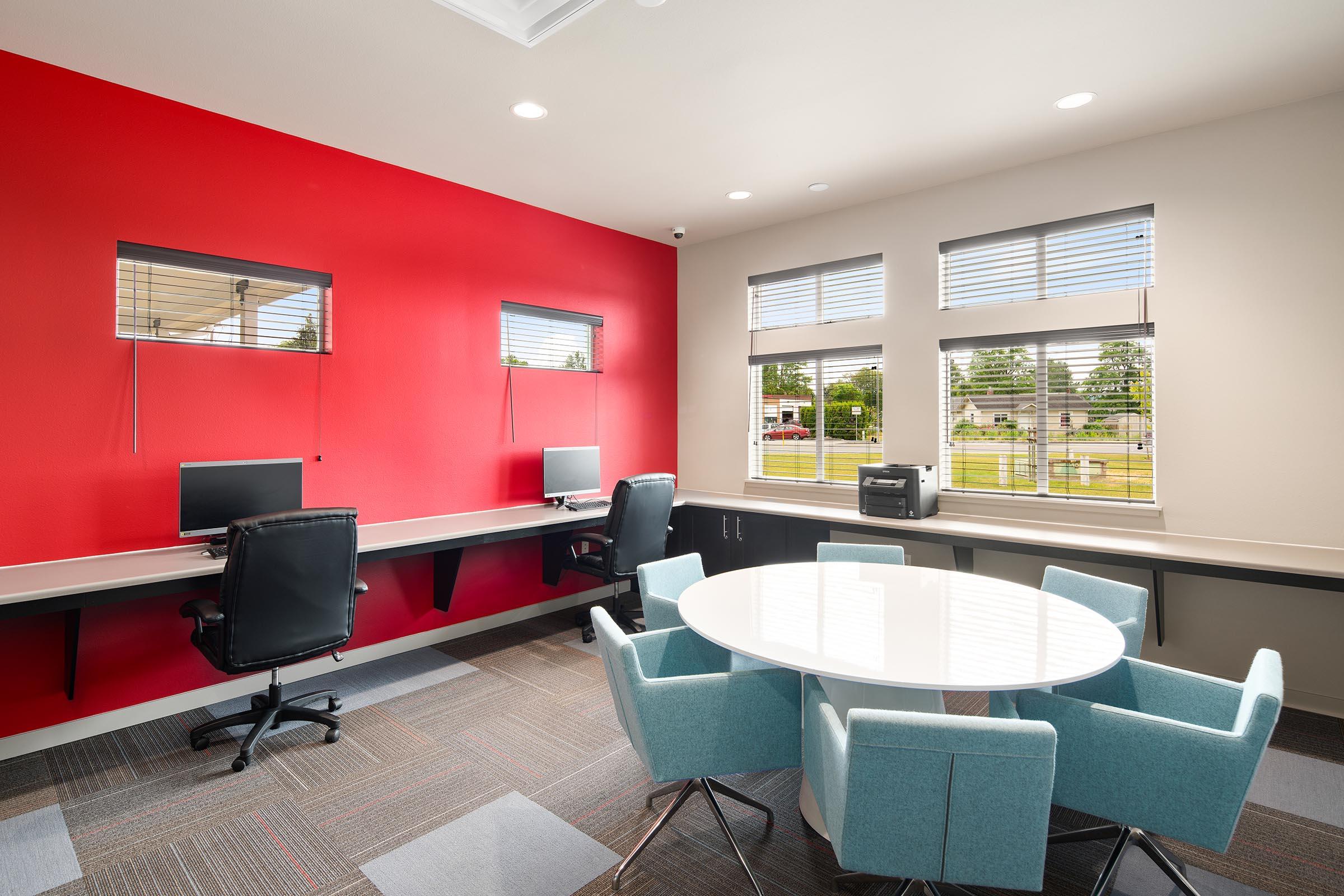A modern office space featuring two computer workstations with black chairs against a red wall. There is a round white table surrounded by light blue chairs in the center, and large windows with blinds allow natural light. The floor is covered with a gray carpet.