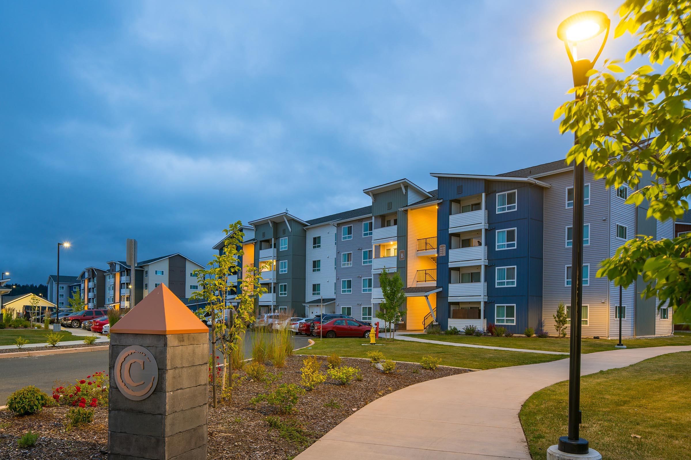 A well-lit apartment complex at dusk features modern architecture with colorful accents. The walkway includes landscaped areas and decorative lighting, leading to the entrance marked by a stone monument. Cars are parked nearby, enhancing the residential atmosphere.