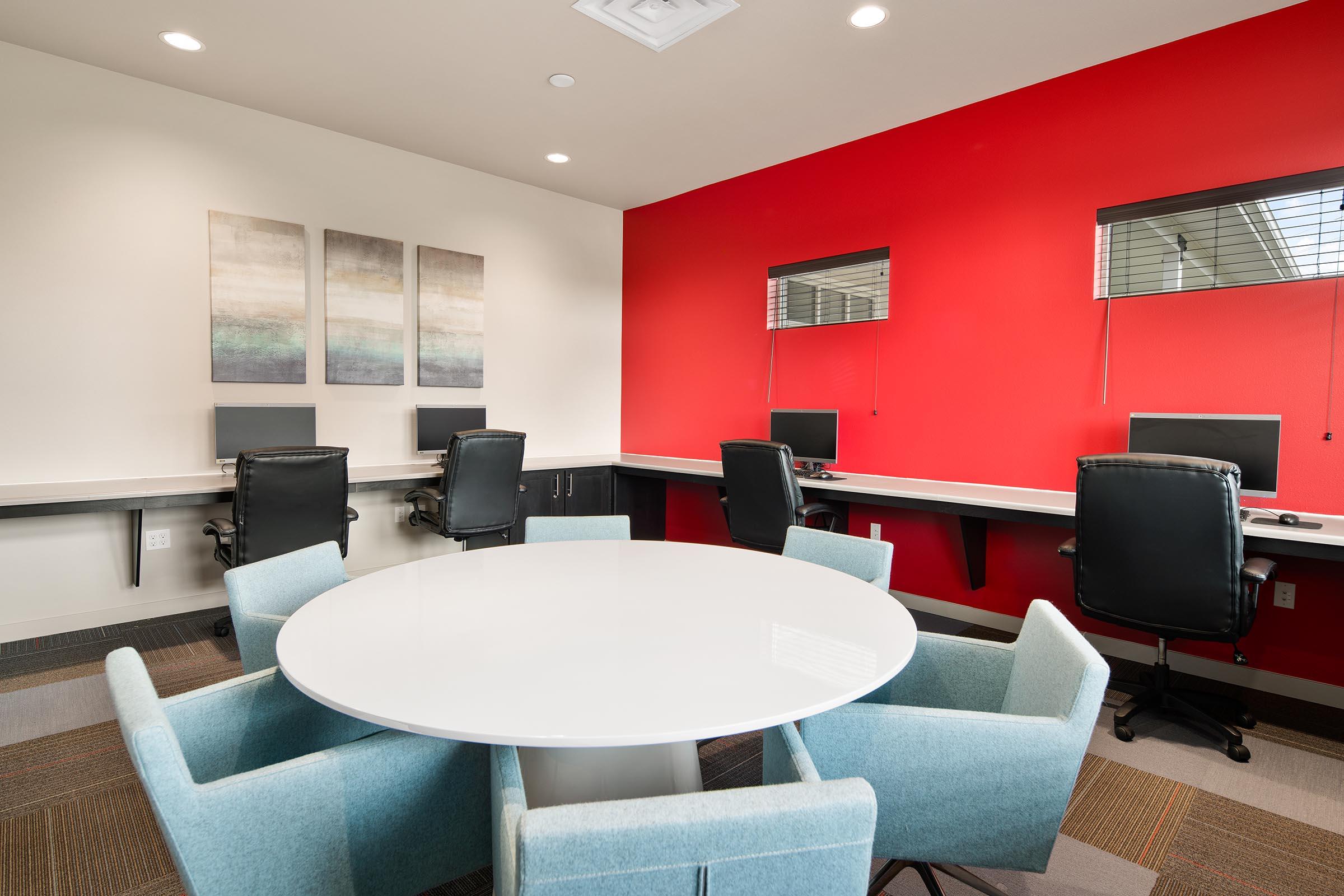 A modern office space featuring a circular white table surrounded by light blue chairs, with two workstations along the wall equipped with computers. The background has a bold red wall and abstract artwork, creating a vibrant yet professional atmosphere.