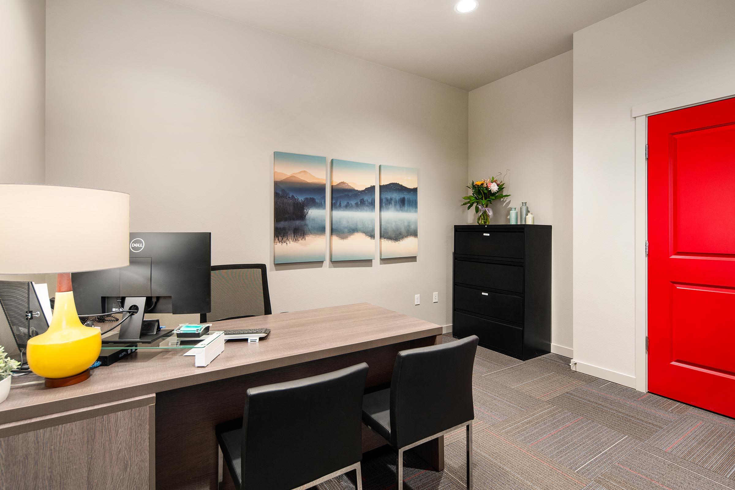 A modern office space featuring a wooden desk with a computer, two black chairs, a decorative lamp, and a filing cabinet. The wall displays a triptych landscape artwork of mountains and a lake. A bright red door adds a pop of color, while a small vase with flowers is on the desk. Soft lighting enhances the inviting atmosphere.