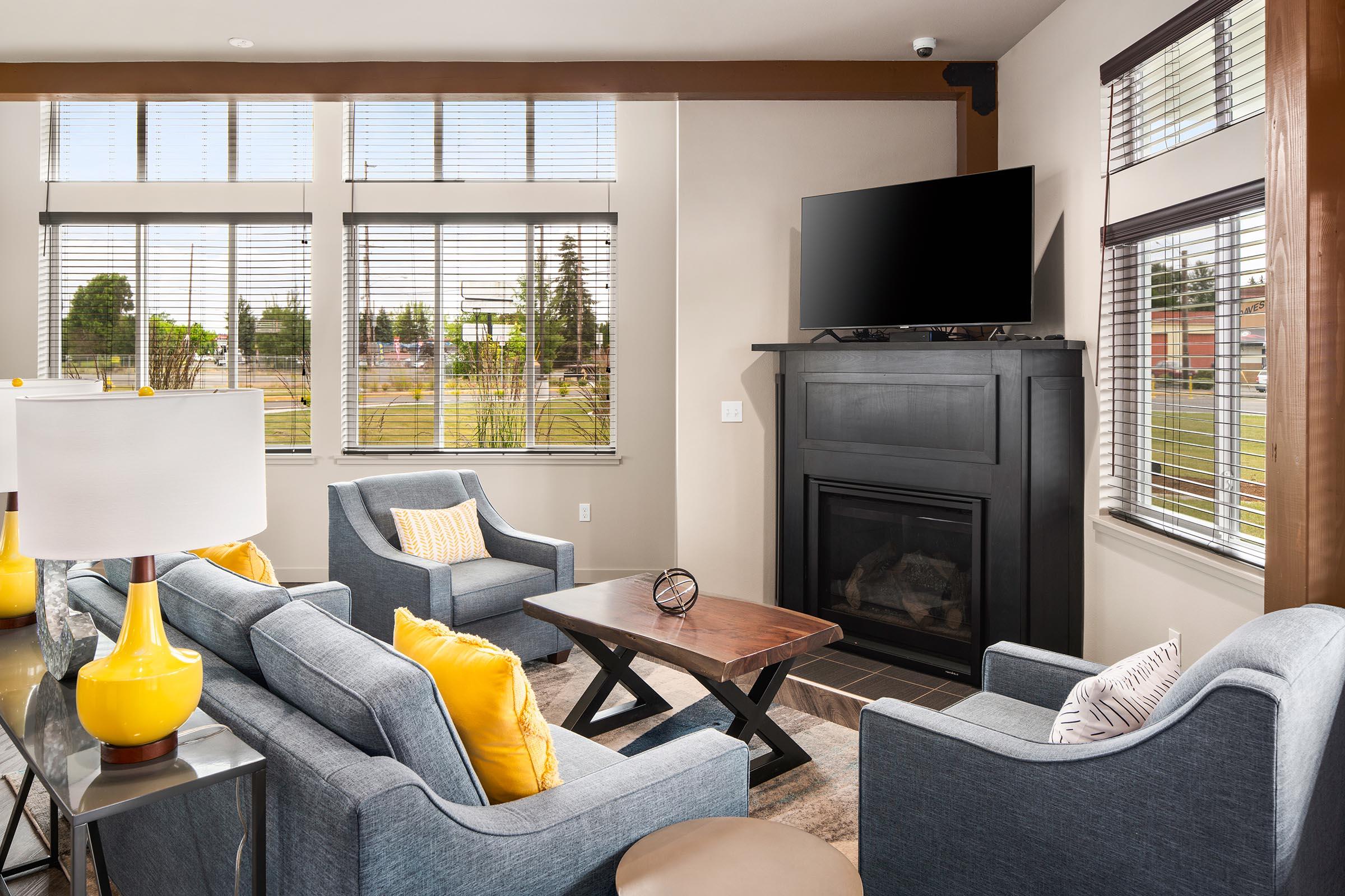 A bright and cozy living room featuring a modern design. There are two light blue sofas with yellow cushions, a dark wooden coffee table, and a stylish lamp on a side table. A flat-screen TV is mounted above a black fireplace, and large windows allow natural light to fill the space, showcasing an outdoor view.