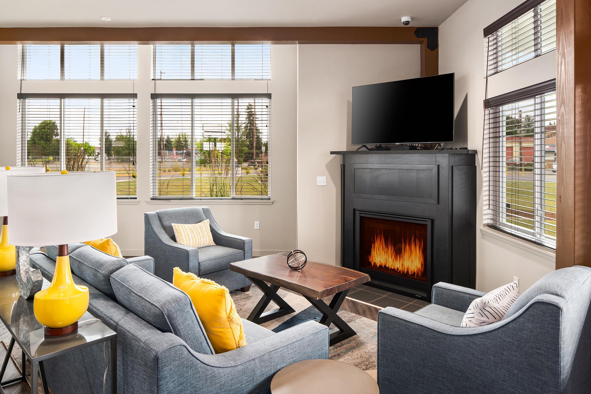 A cozy living room featuring a gray sofa and two blue armchairs with yellow pillows. There's a wooden coffee table in the center, and a modern fireplace beneath a wall-mounted television. Large windows provide ample natural light, and light-colored walls enhance the space's inviting atmosphere.