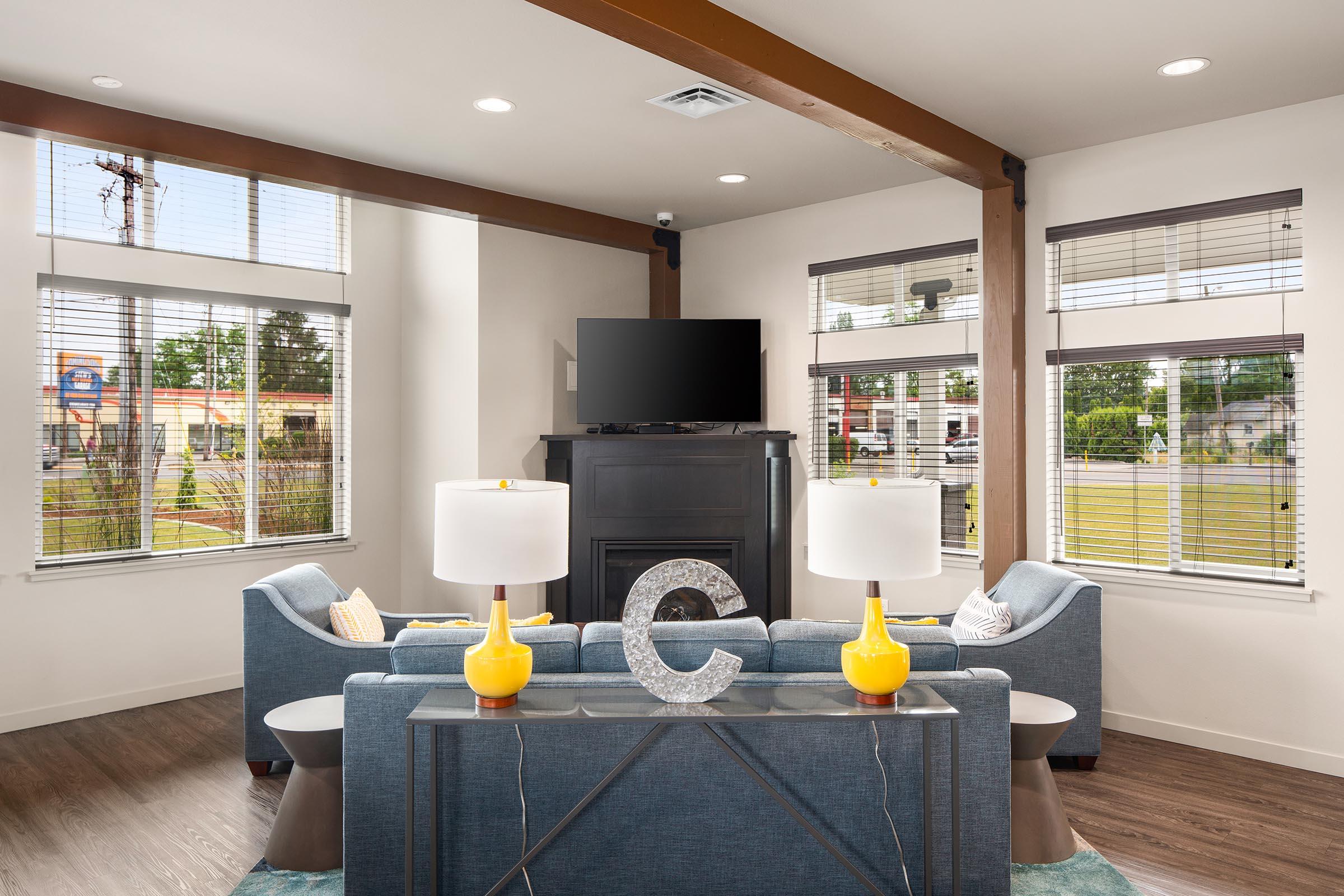 Modern living room featuring a blue sofa set, two yellow lamps on a glass table, and a dark fireplace. A flat-screen TV is mounted on the wall. The space has large windows with blinds, allowing natural light, and a view of a green area outside. The decor is contemporary and inviting.