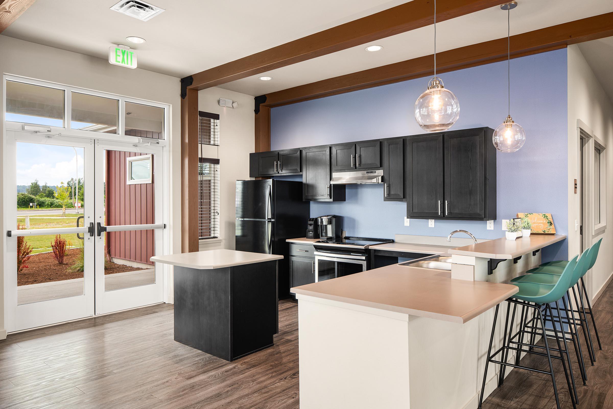 Modern kitchen interior with an open layout. Features black appliances, a white island, and a wooden bar counter with high stools. Natural light enters through large windows, and the decor includes a light blue accent wall and wooden beams on the ceiling.