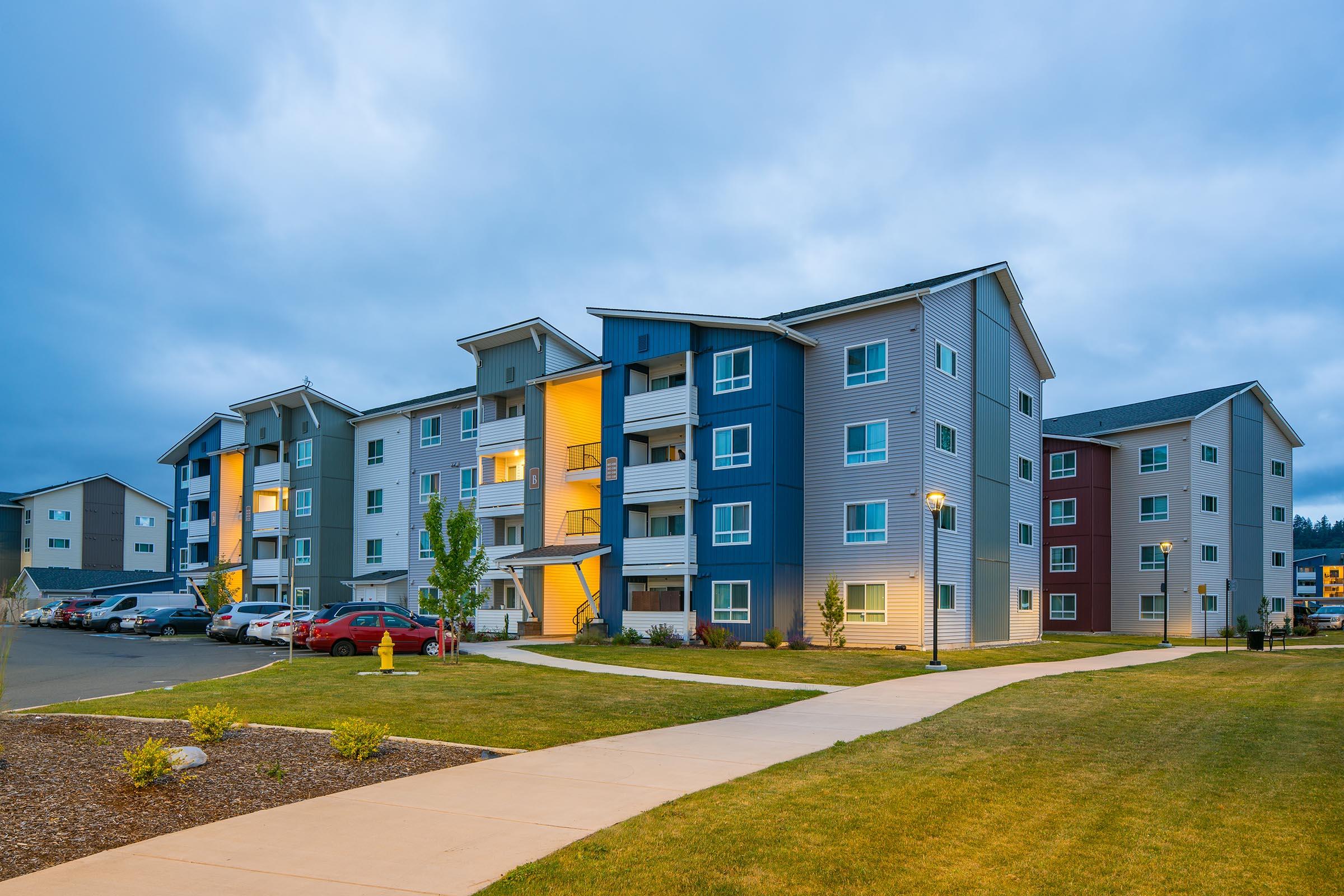 A modern, multi-story apartment building with a mix of blue, gray, and red exteriors. The scene is set during dusk, with some lights on in the apartments. A pathway runs in front, leading to the entrance, and there are parked cars and landscaped areas around the building.