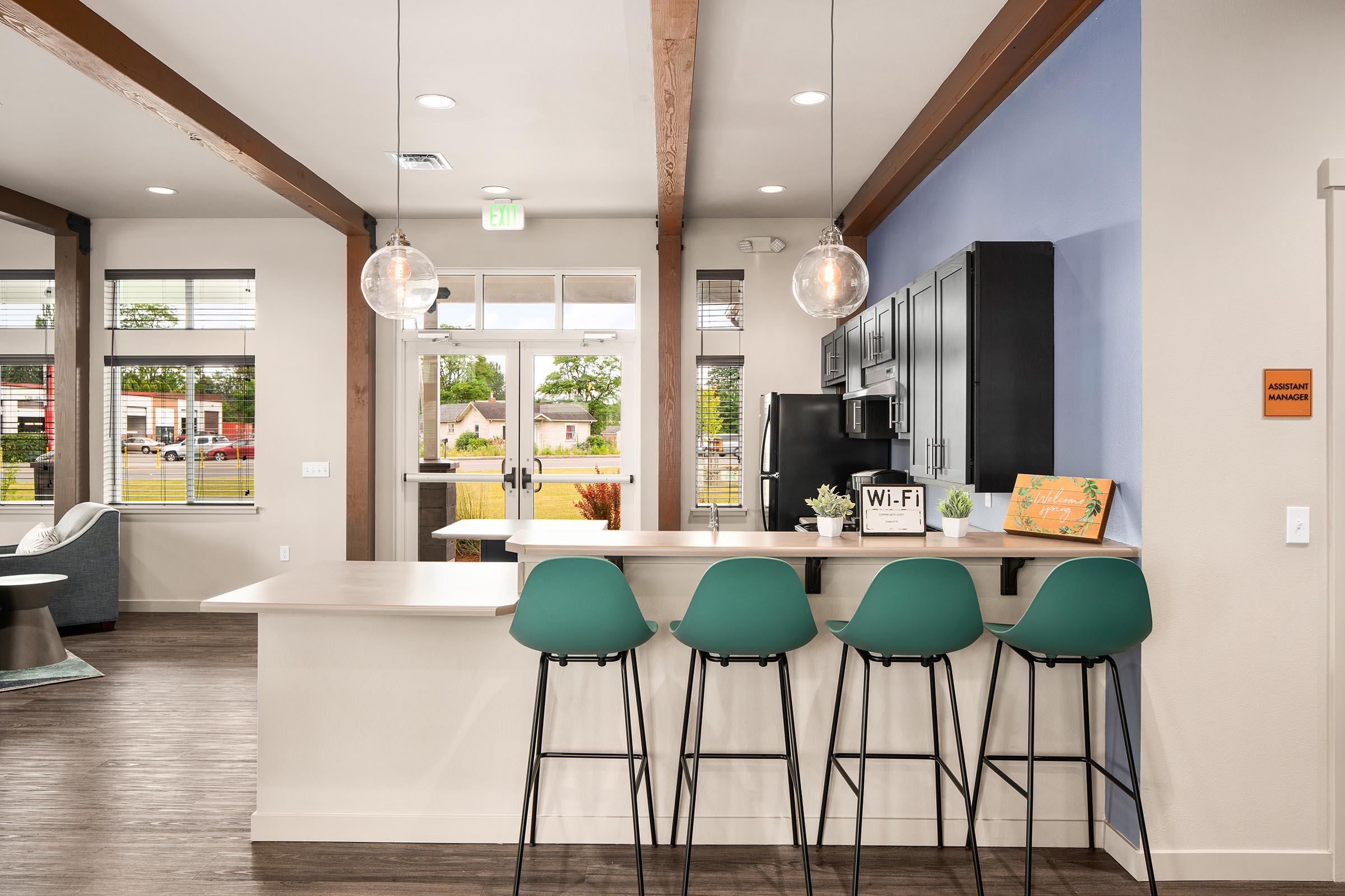 Modern kitchen and lounge area featuring a sleek bar with green stools, a countertop, and contemporary cabinets. Bright lighting fixtures hang above the bar, and large windows allow natural light to fill the space. A welcoming atmosphere is created with plants and a sign indicating Wi-Fi availability.