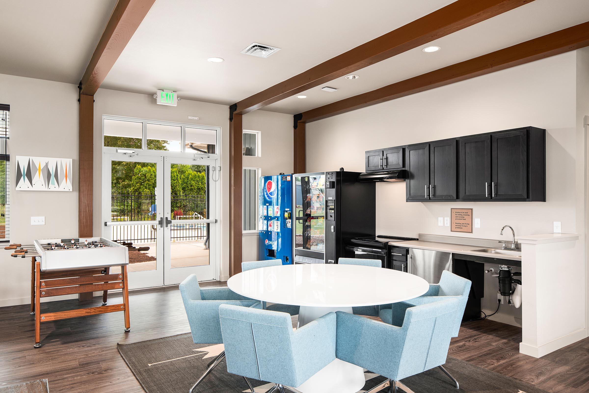 Modern common area featuring a round white table with light blue chairs, a black kitchenette with appliances, a vending machine, and a foosball table. Large windows allow natural light, enhancing the spacious feel with a contemporary design and wooden beams on the ceiling.