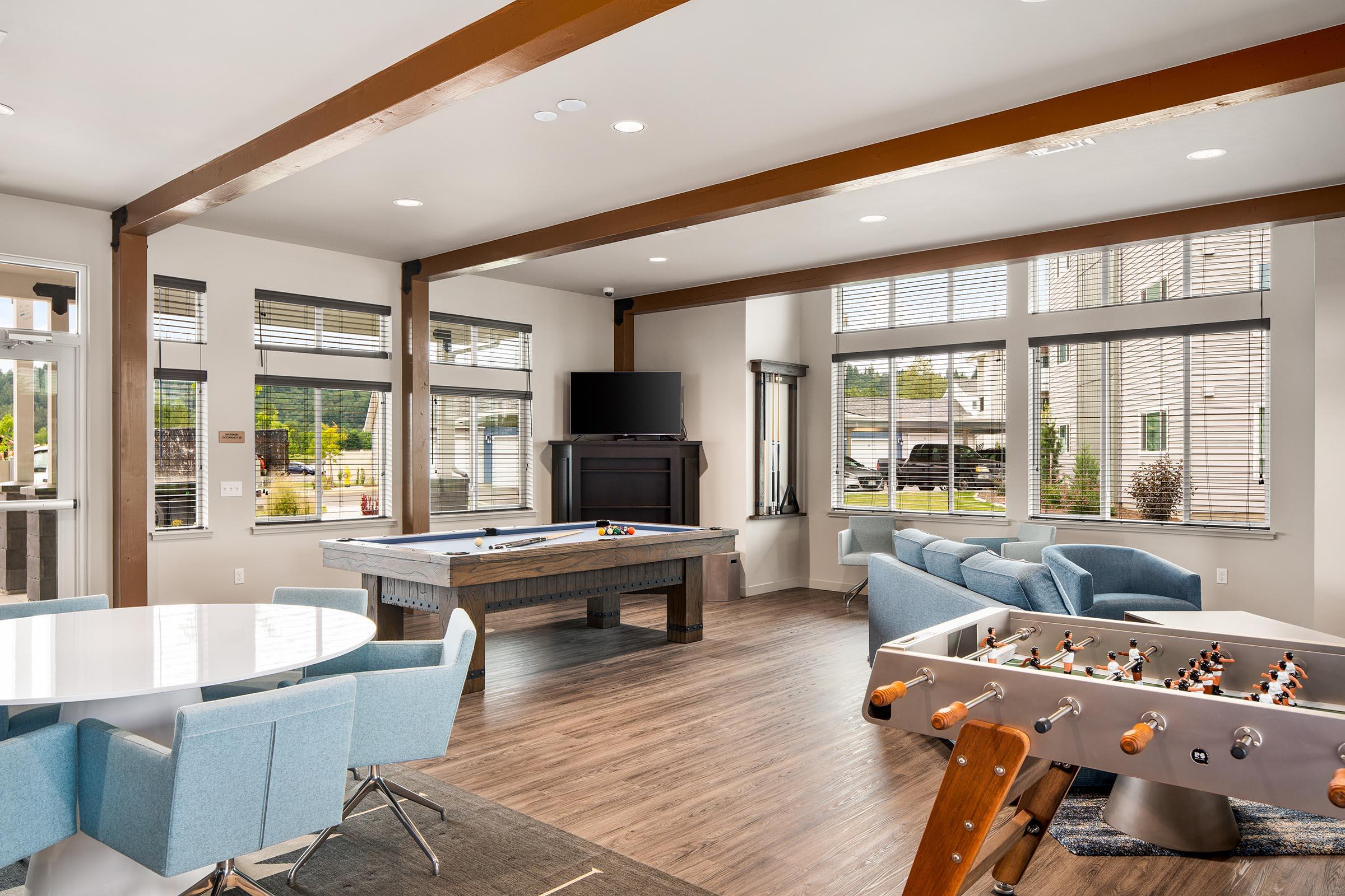 A modern lounge area featuring a pool table and a foosball table. Brightly lit with large windows, the space includes a round dining table with blue chairs and a comfortable seating arrangement. Decorative elements and wooden beams add a contemporary touch to the interior.