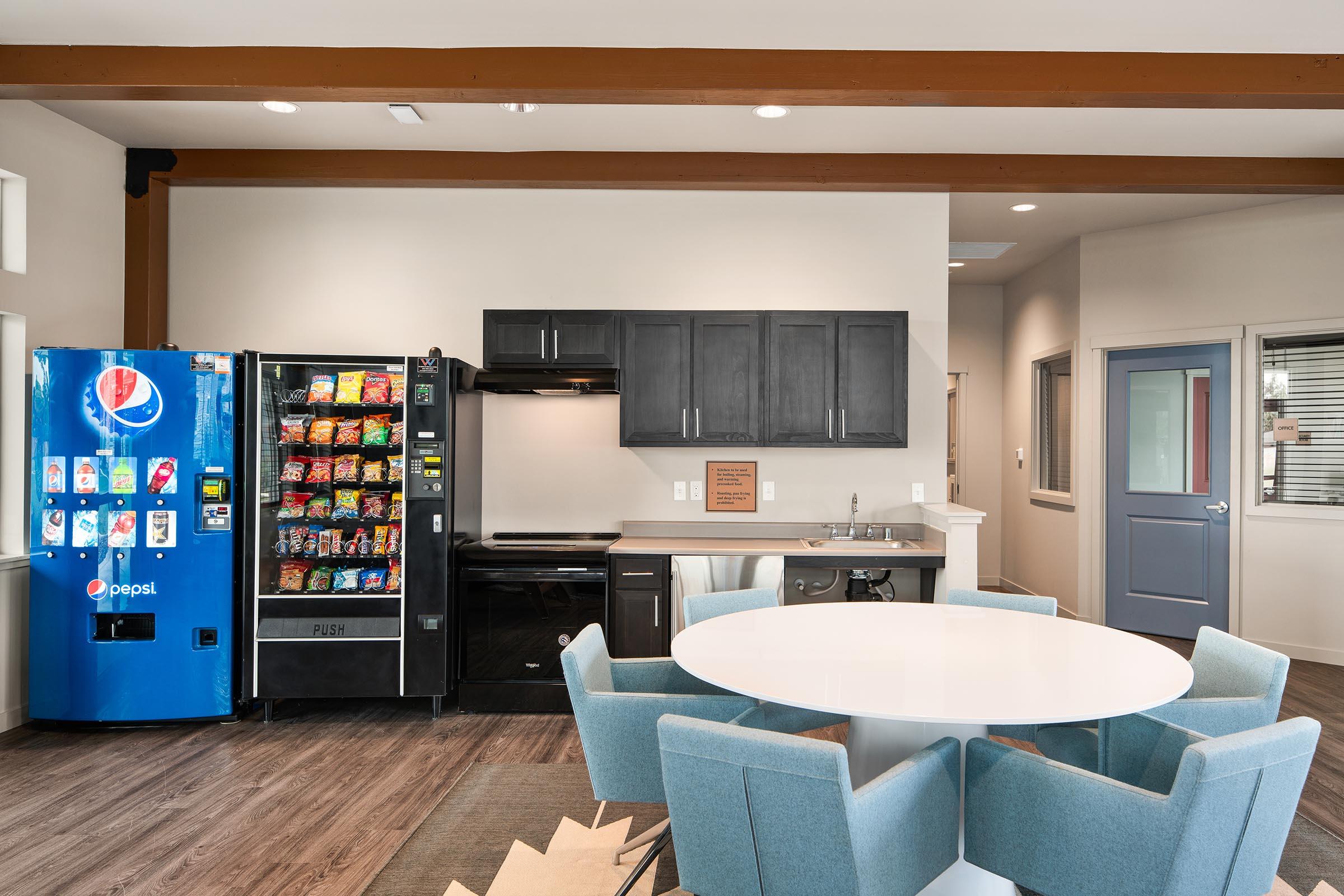 A modern break room featuring a round white table surrounded by light blue chairs. On one side, there’s a vending machine stocked with snacks and beverages, and next to it, a black kitchen area with an oven and countertop. Large windows allow natural light in, enhancing the inviting atmosphere.