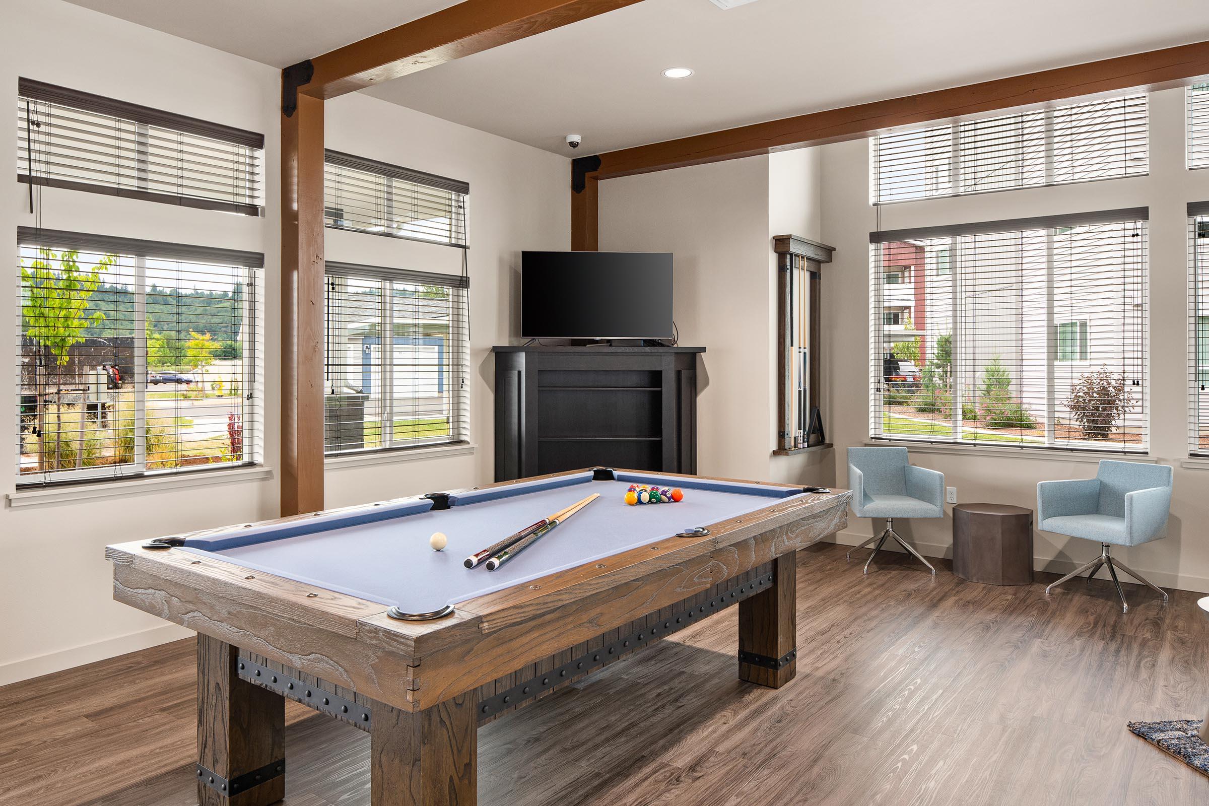 A modern game room featuring a pool table with cues and balls, large windows allowing natural light, and contemporary furniture including two light blue chairs and a small side table. A flat-screen TV is mounted on the wall. The design has wooden beams and a warm color palette.
