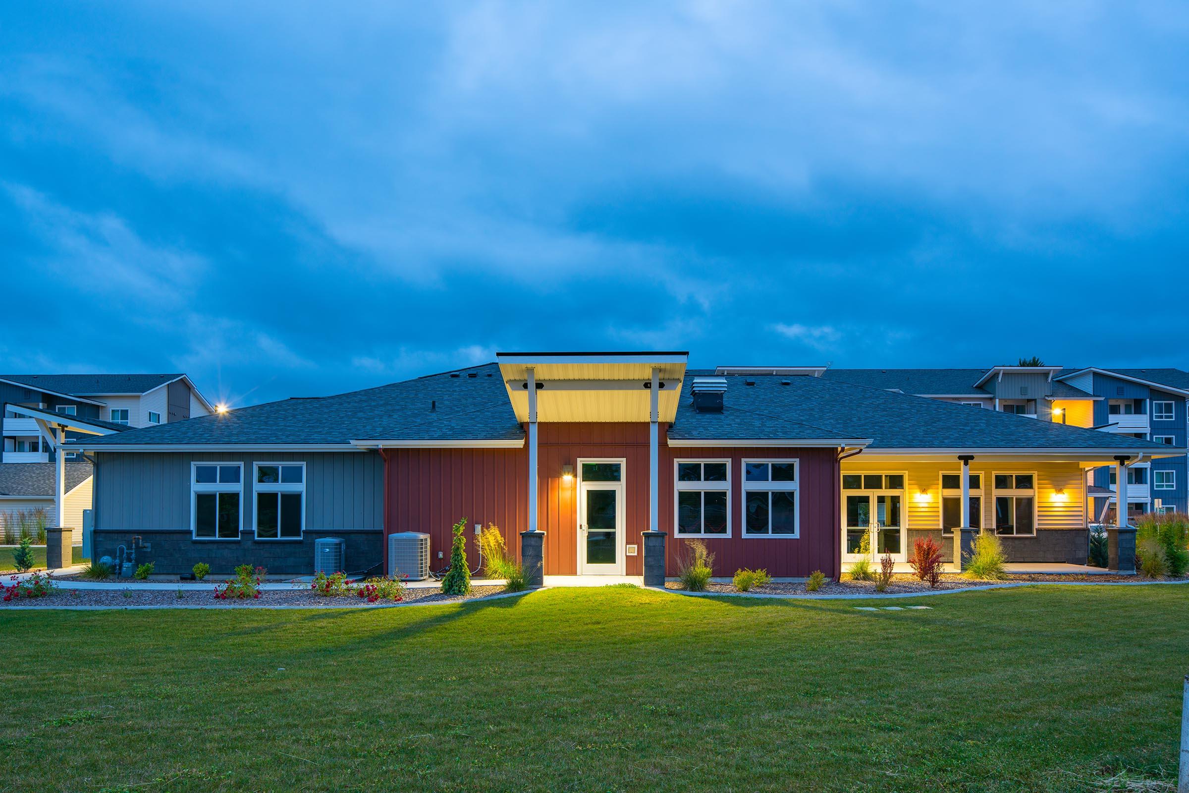 A modern multi-unit residential building at twilight, featuring a blend of red, blue, and yellow siding. The structure is well-lit, highlighting landscaped gardens and a green lawn in the foreground. Cloudy skies create a serene evening atmosphere, with additional buildings visible in the background.