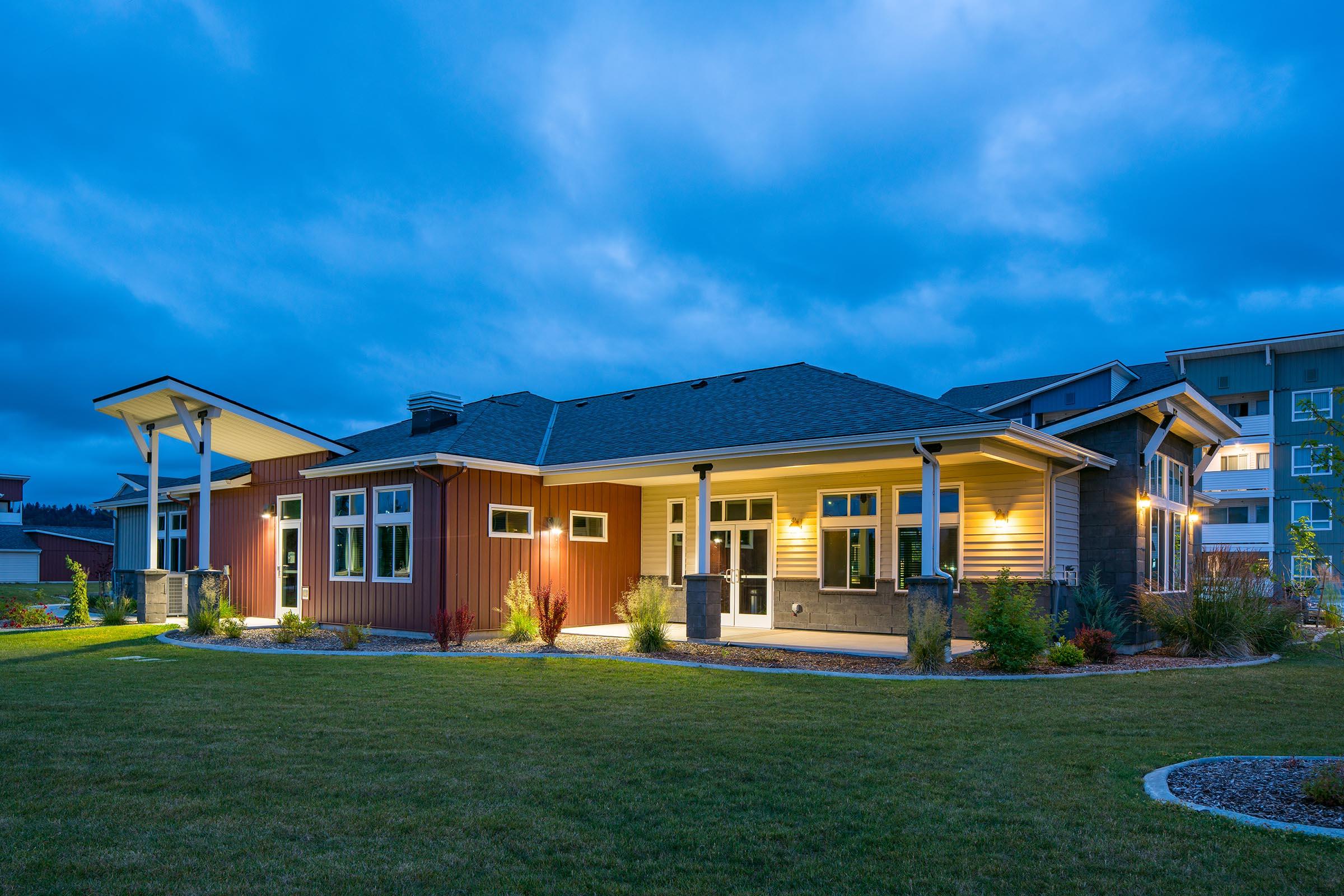 A modern house at dusk, showcasing a combination of dark red and light brown siding. Soft lighting highlights the architecture, with landscaping featuring green grass and planted shrubs. The scene has a calm atmosphere, with a cloudy blue sky in the background.