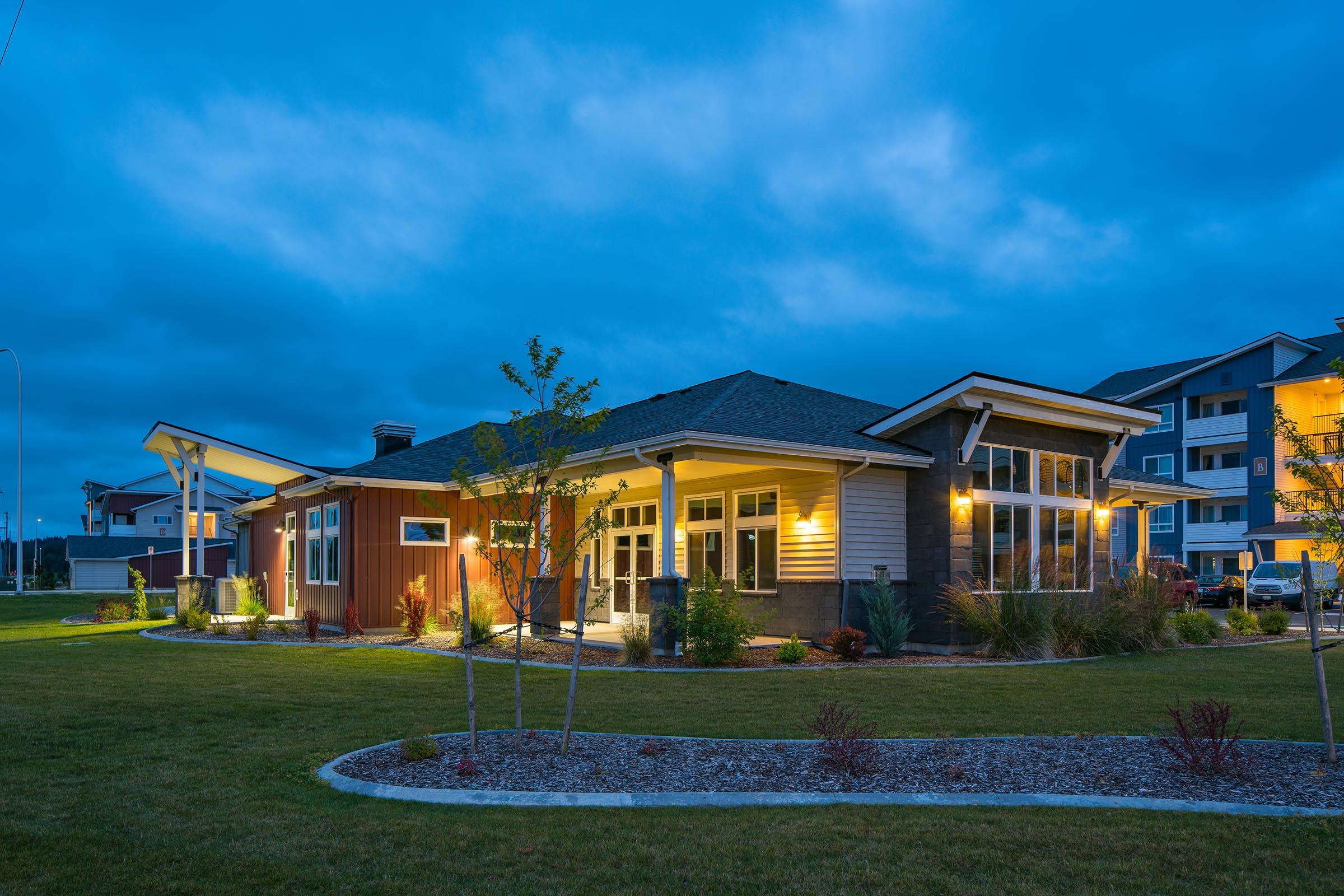 A well-lit modern house at dusk, featuring a mix of wood and stone materials. Surrounded by landscaped grass and small trees, with a cloudy blue sky overhead. The house showcases large windows and a welcoming entrance. In the background, an apartment building is partially visible.