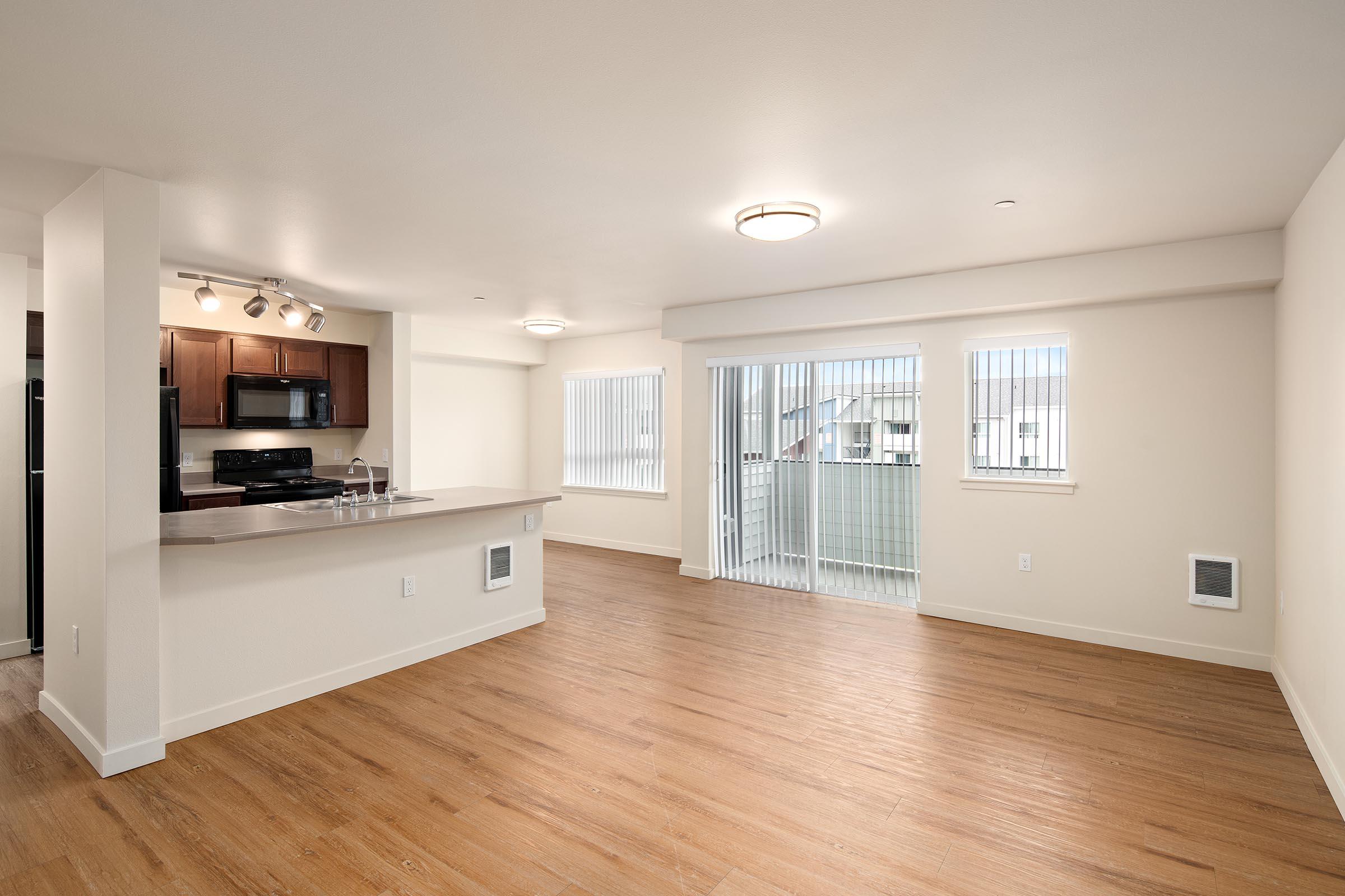 Spacious modern apartment interior featuring an open layout with hardwood floors, a kitchen area equipped with dark wood cabinets and stainless steel appliances, and large windows allowing natural light. Patio doors lead to a balcony, providing outdoor access.