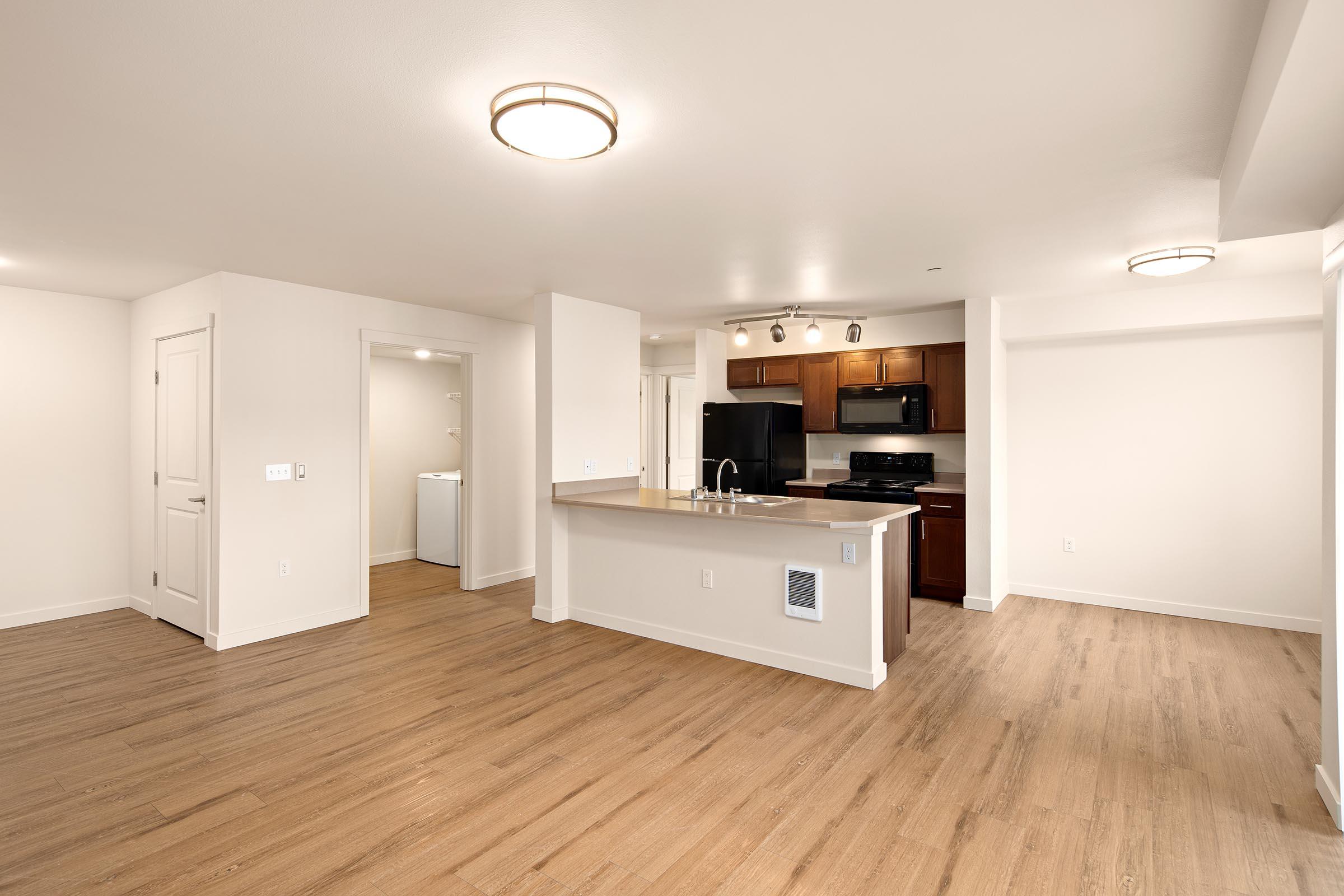 A modern, spacious apartment interior featuring an open layout. The kitchen has dark wood cabinets and black appliances, while the living area has light-colored walls and wood flooring. A laundry area is visible through an open door, and the space is well-lit with ceiling lights.