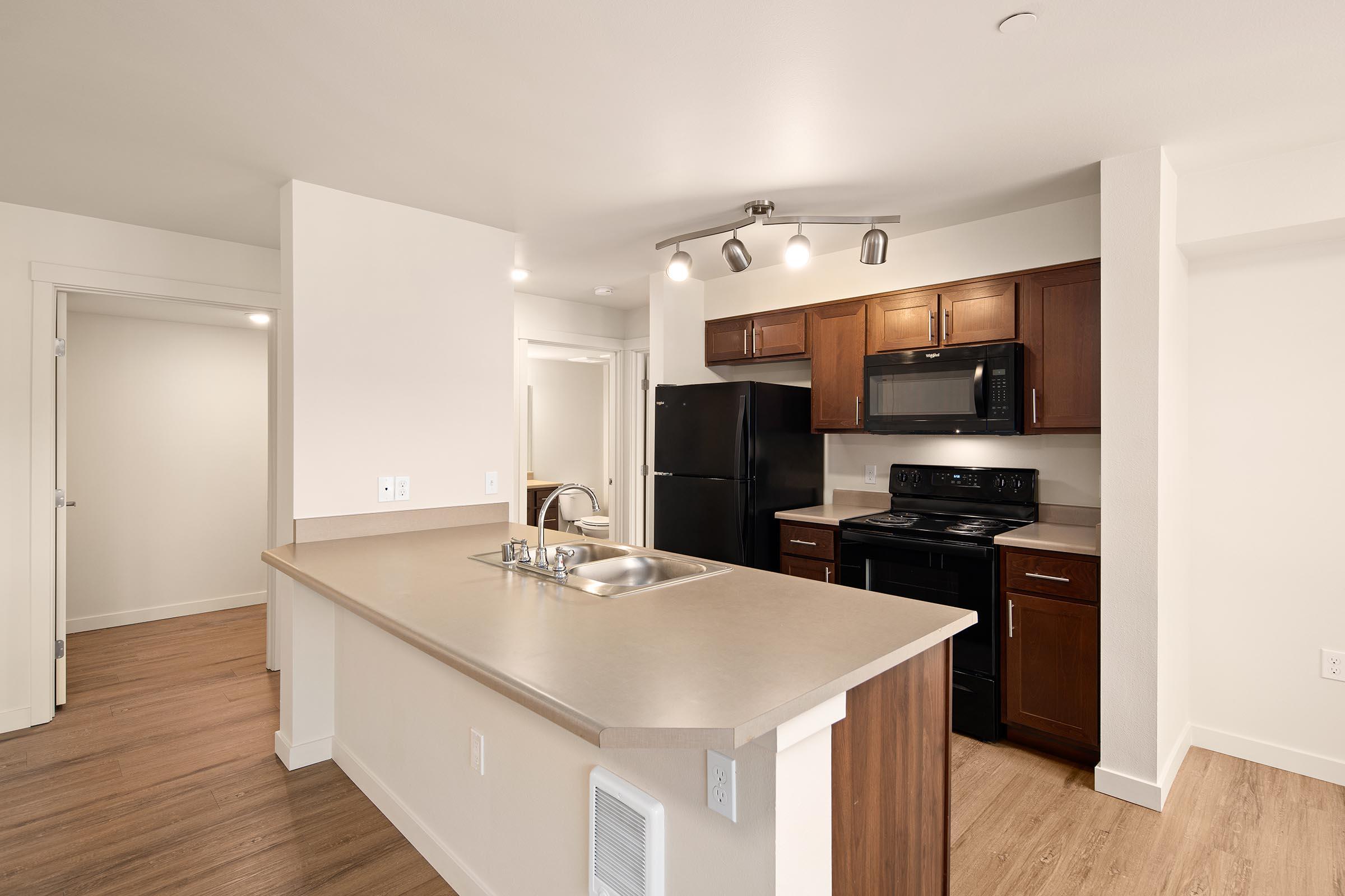 Modern kitchen with a central island, featuring dark wood cabinets, stainless steel appliances including a microwave and refrigerator, and a neutral countertop. The space has warm wood flooring and is well-lit with overhead lights. A doorway leads to another room in the background.