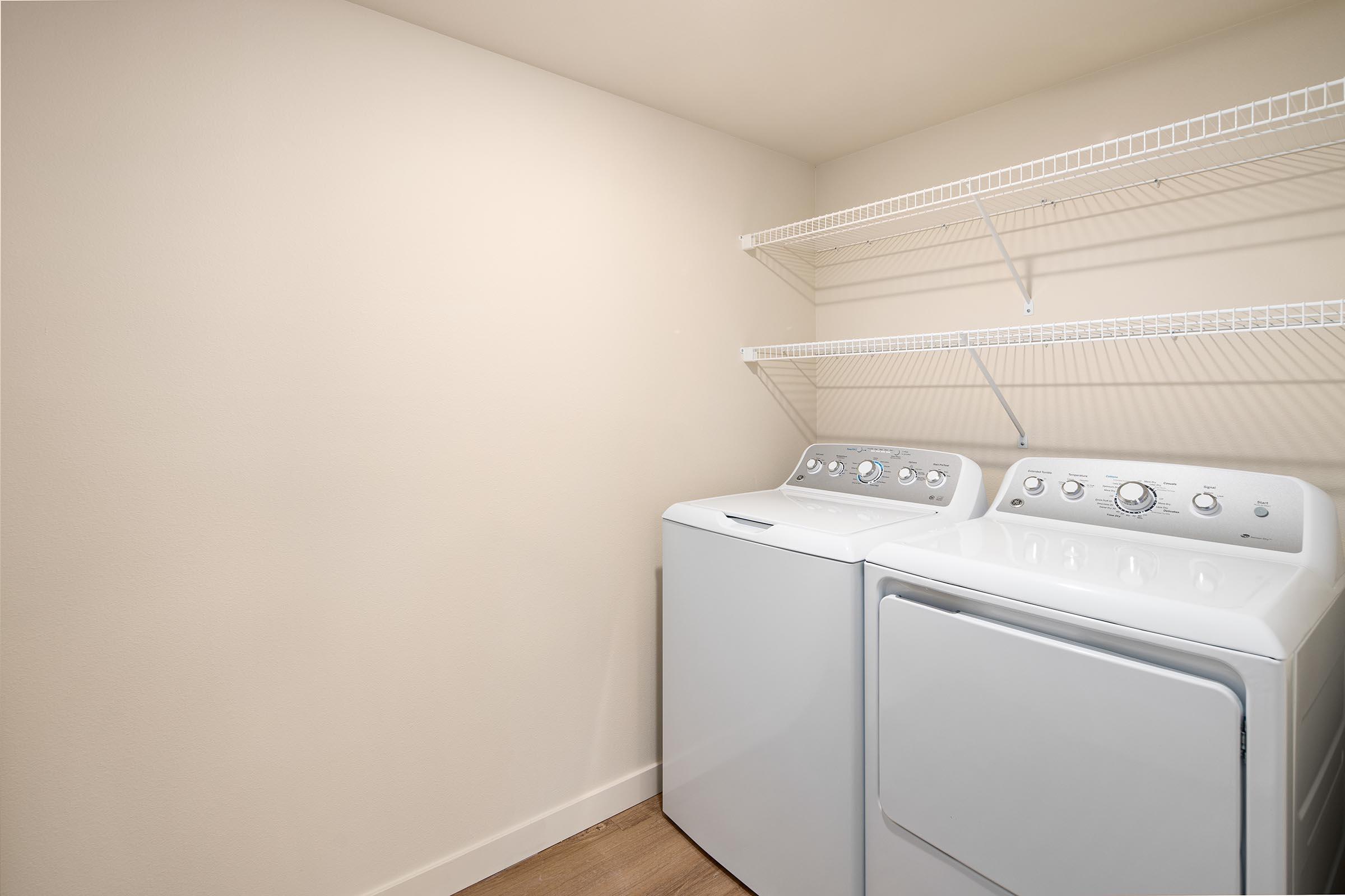 A clean, minimal laundry room featuring a white washing machine and dryer side by side, with a wall-mounted shelf above. The walls are painted in a light neutral color, and the floor is made of light wood, creating a tidy and modern look.