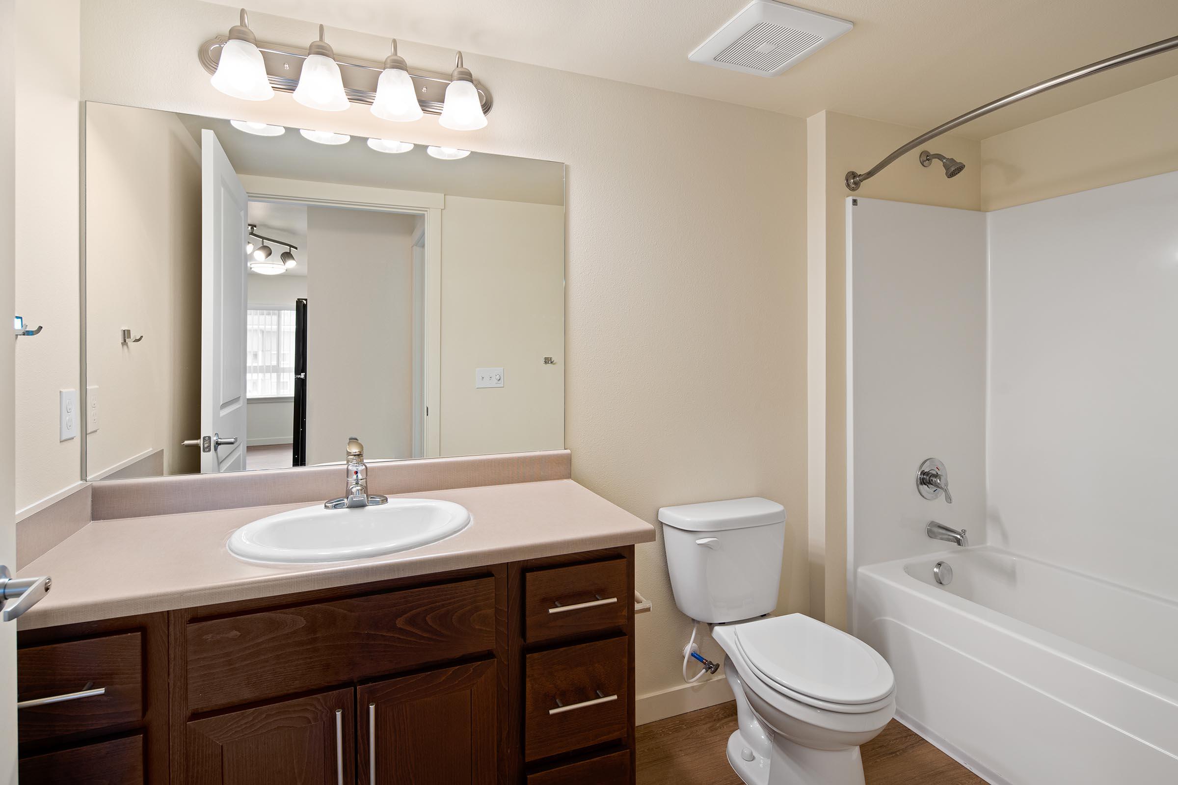 A clean and modern bathroom featuring a wall-mounted mirror above a sink with a single faucet. There is a toilet, a bathtub with shower curtain, and light fixtures above the mirror. The walls are painted a light color, and the flooring is a warm wood tone, creating a spacious and inviting atmosphere.