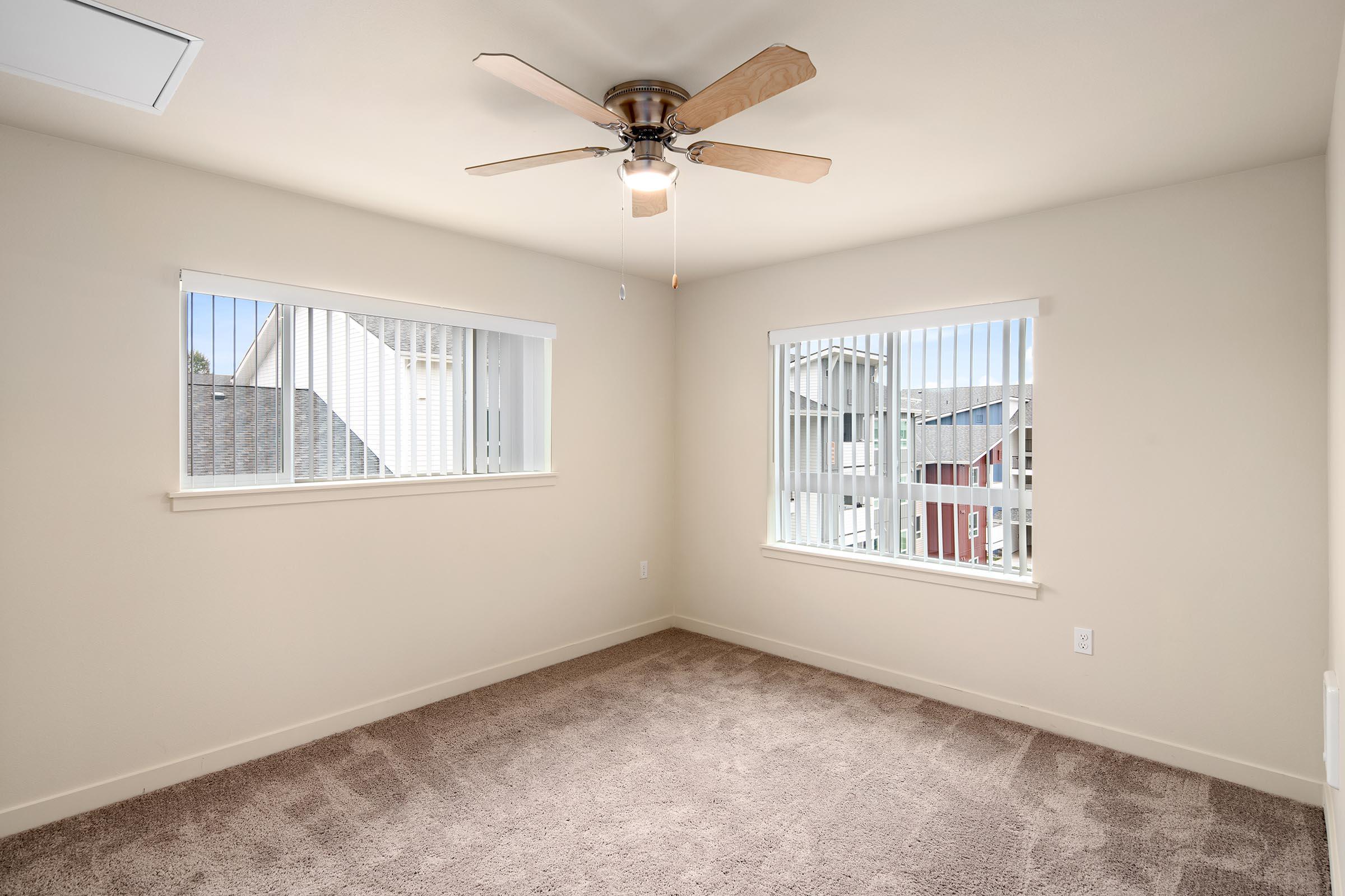 A bright, empty room featuring two windows with vertical blinds and a ceiling fan. The walls are painted a light color, and the floor is carpeted in a neutral shade. The room is organized and inviting, showcasing potential for various uses such as a bedroom or a home office.