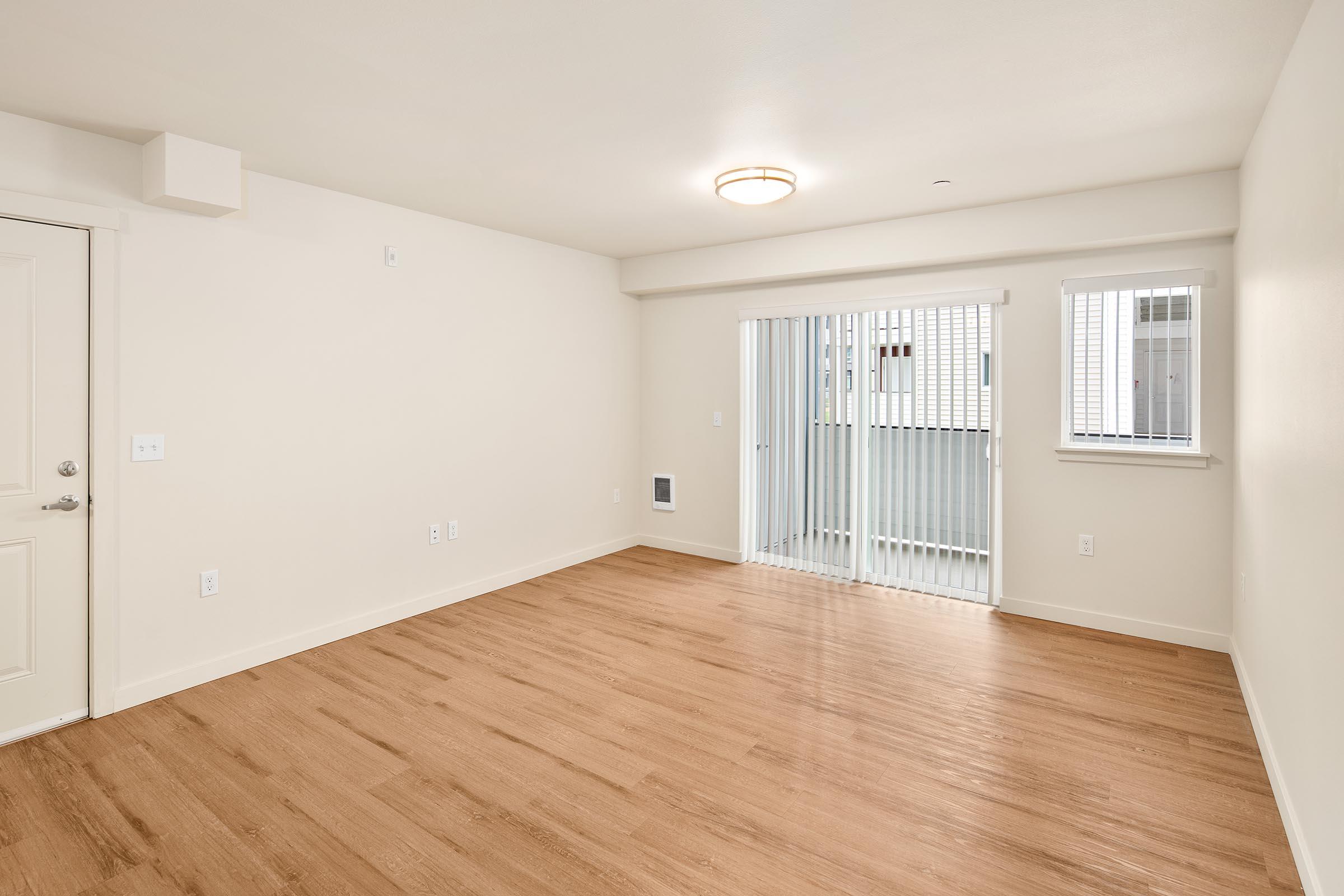 Empty interior of a modern apartment with light-colored walls and a wood-like floor. Large window with vertical blinds and a sliding door leading to a small balcony. Room features minimal decor, creating a spacious and inviting atmosphere.