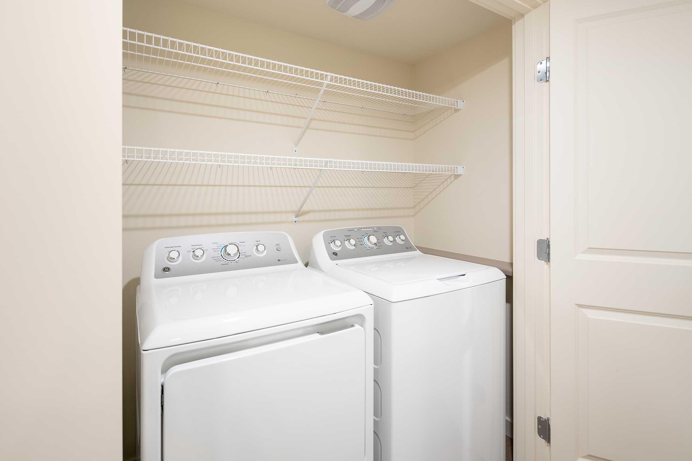 A laundry room featuring a stacked washer and dryer unit. Above them, there are two wire shelves for storage. The walls are a light color, and the door to the room is partially open, revealing the interior space.