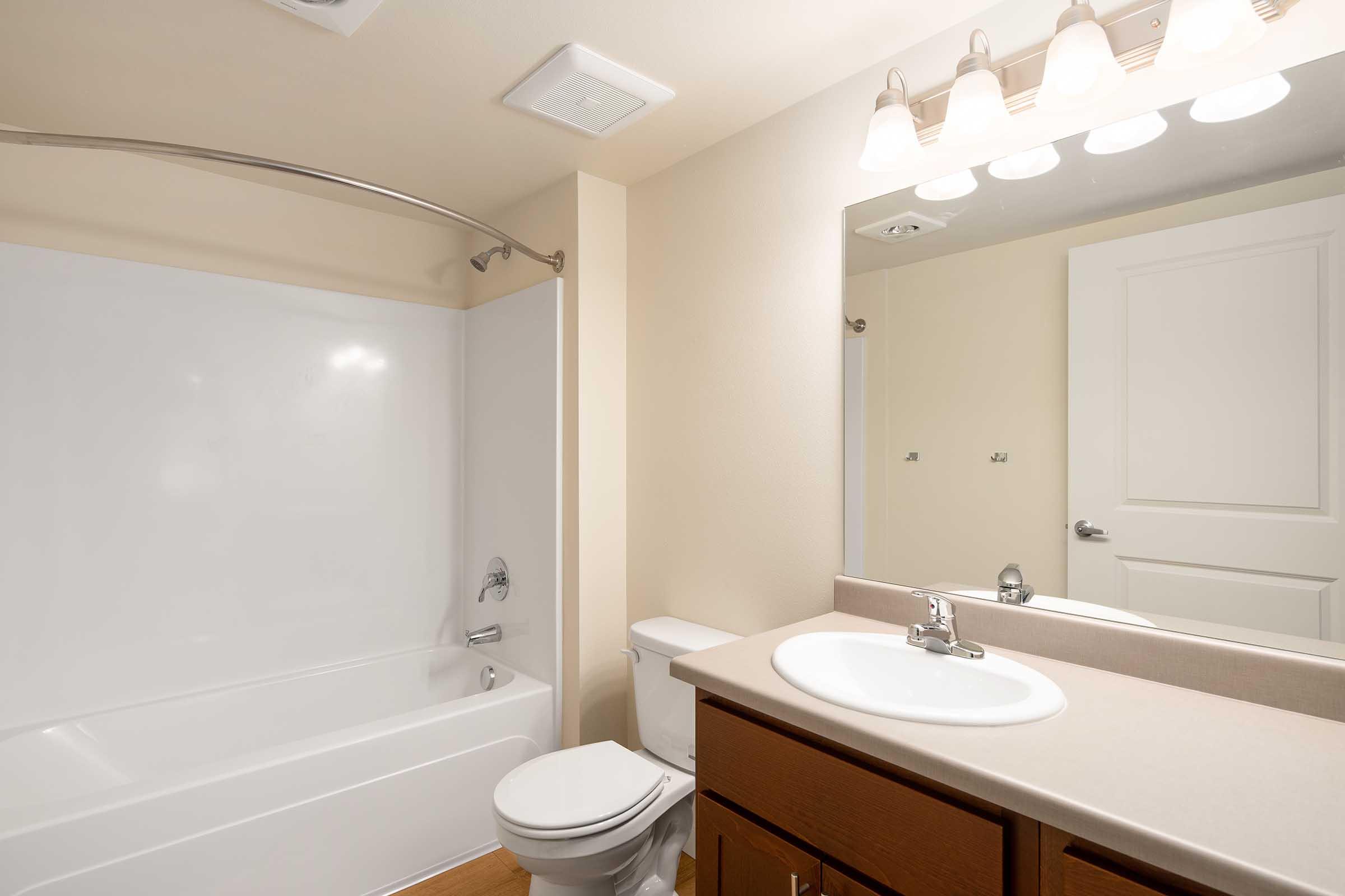A clean and well-lit bathroom featuring a white bathtub with a curved shower rod, a sink with a modern faucet, a wall-mounted mirror, a toilet, and beige walls. The lighting is soft, providing a warm atmosphere. The flooring is wood-like, adding to the overall aesthetic.