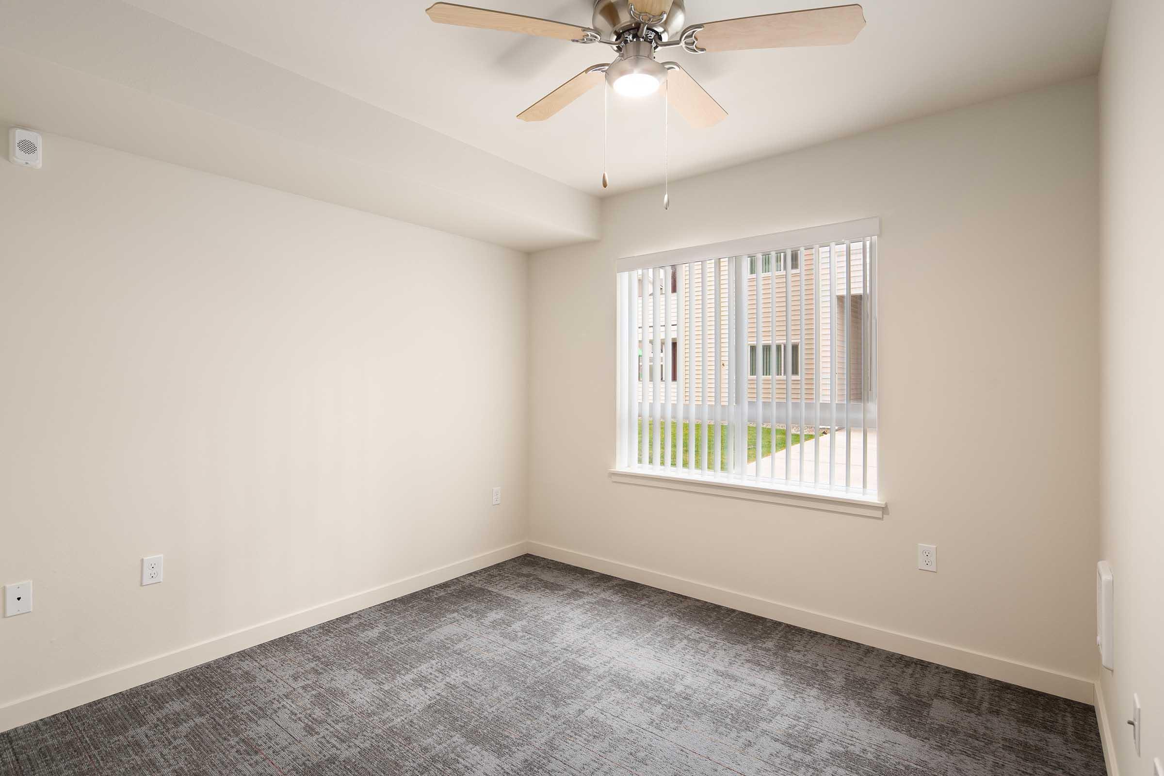 Empty interior room with beige walls and a ceiling fan. There is a window with blinds that lets in natural light, overlooking a green area outside. The floor is covered with gray carpet, and there are electrical outlets on the walls.