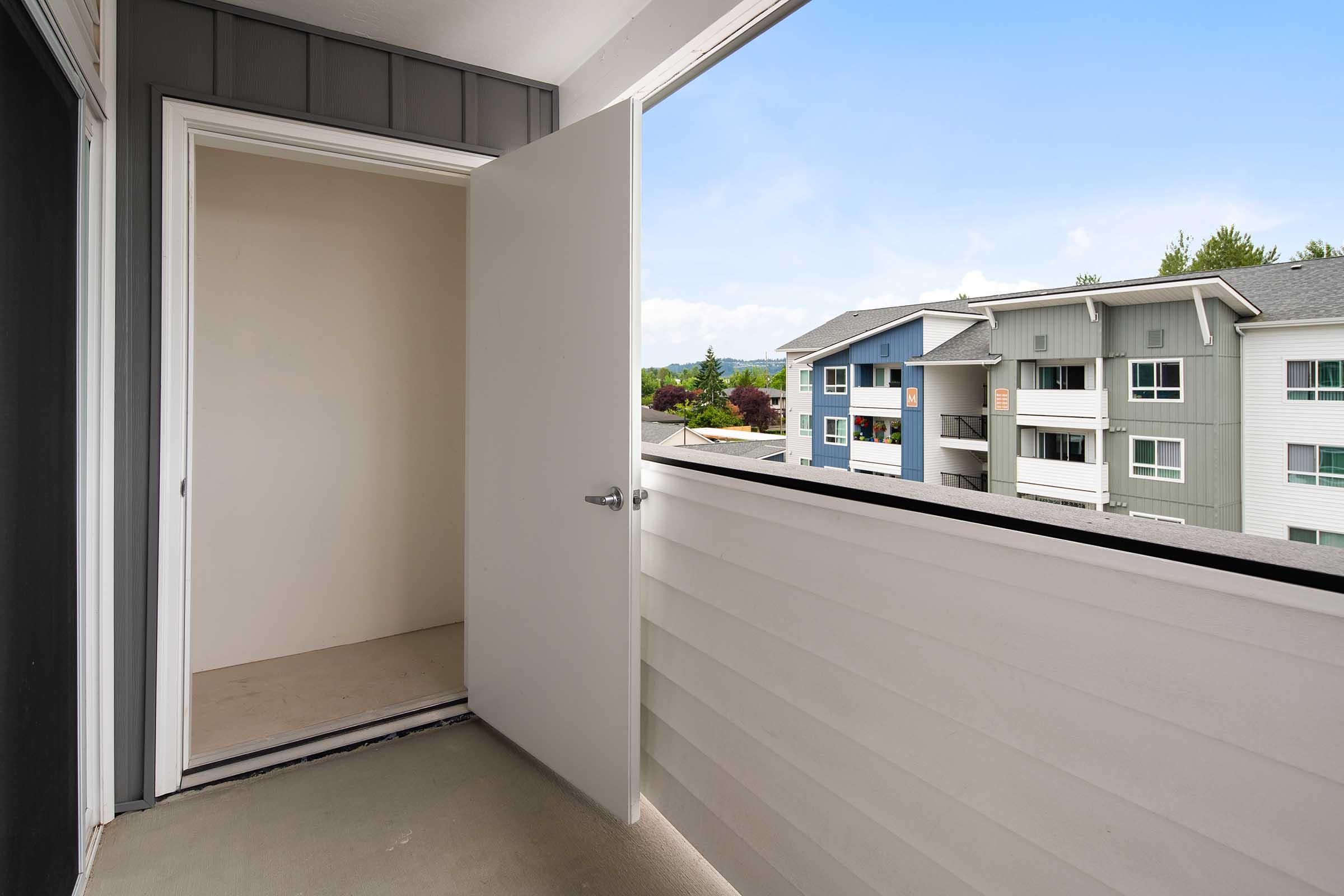 An open door leads to a small balcony overlooking an apartment complex. The balcony has a plain concrete floor, with a view of nearby buildings painted in various colors. The sky is blue with a few clouds, creating a bright and inviting atmosphere.