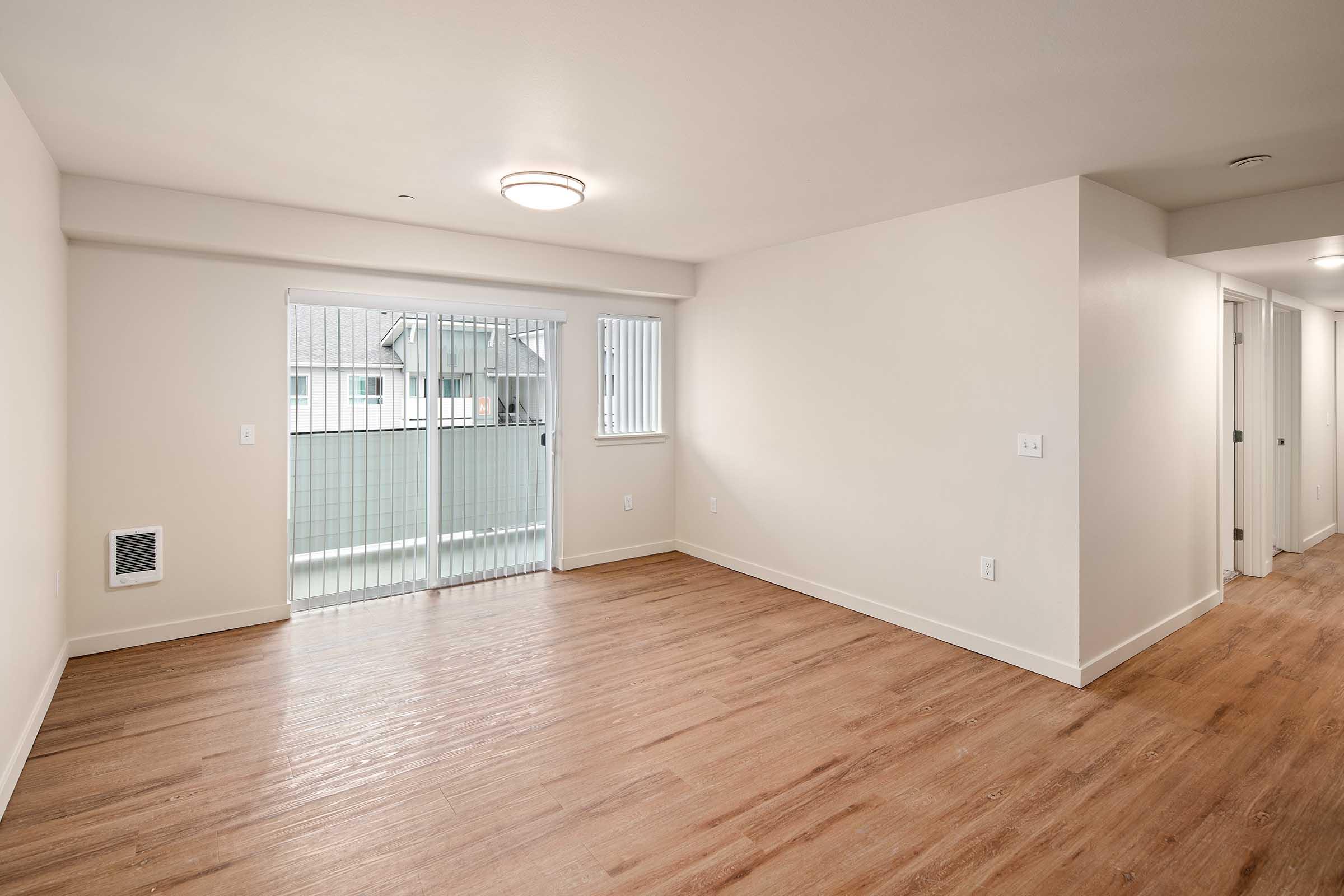 Spacious, empty room with light-colored walls and wooden flooring, featuring a sliding glass door that leads to an outdoor balcony. The room is well-lit, with a ceiling light fixture and a vent on one wall. There are doorways on the right leading to other areas, enhancing the open feel of the space.