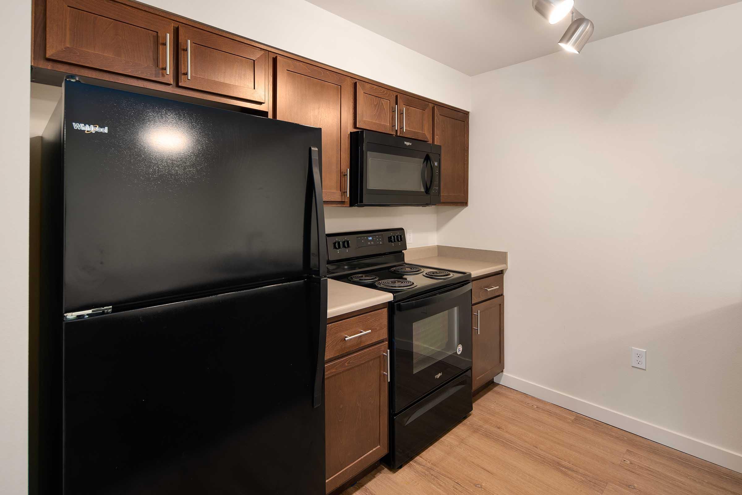 Modern kitchen featuring dark wood cabinets, a black refrigerator, and stainless steel appliances including a microwave and stove. The countertops are light-colored, and there is a neutral wall backdrop. The flooring is wood, creating a warm and inviting atmosphere.